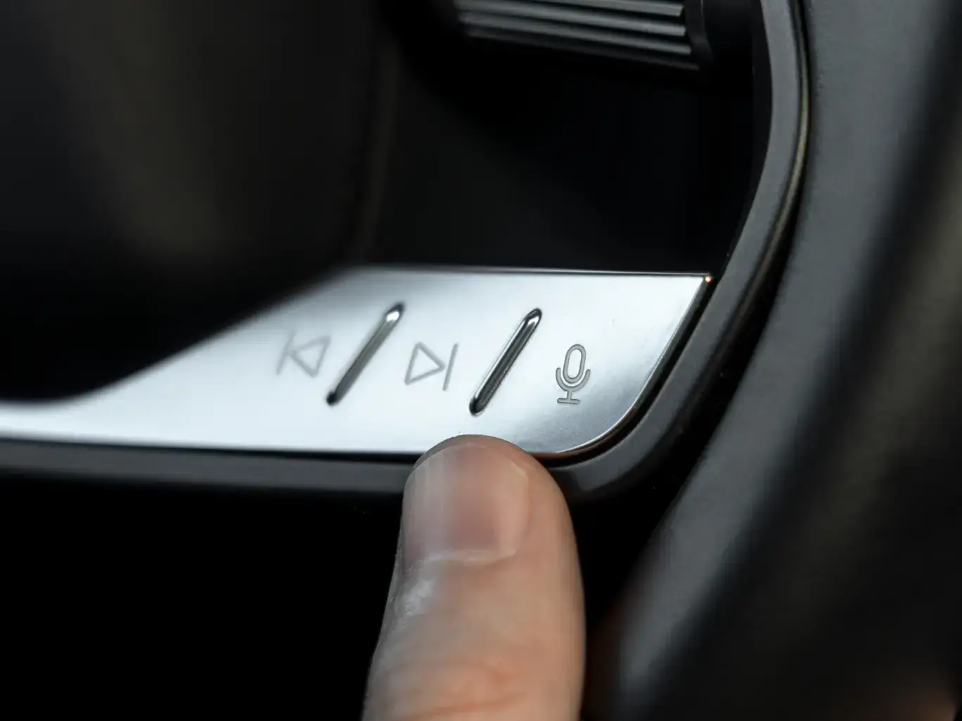 Finger pressing a voice command microphone button on a car's silver media control panel with playback controls.