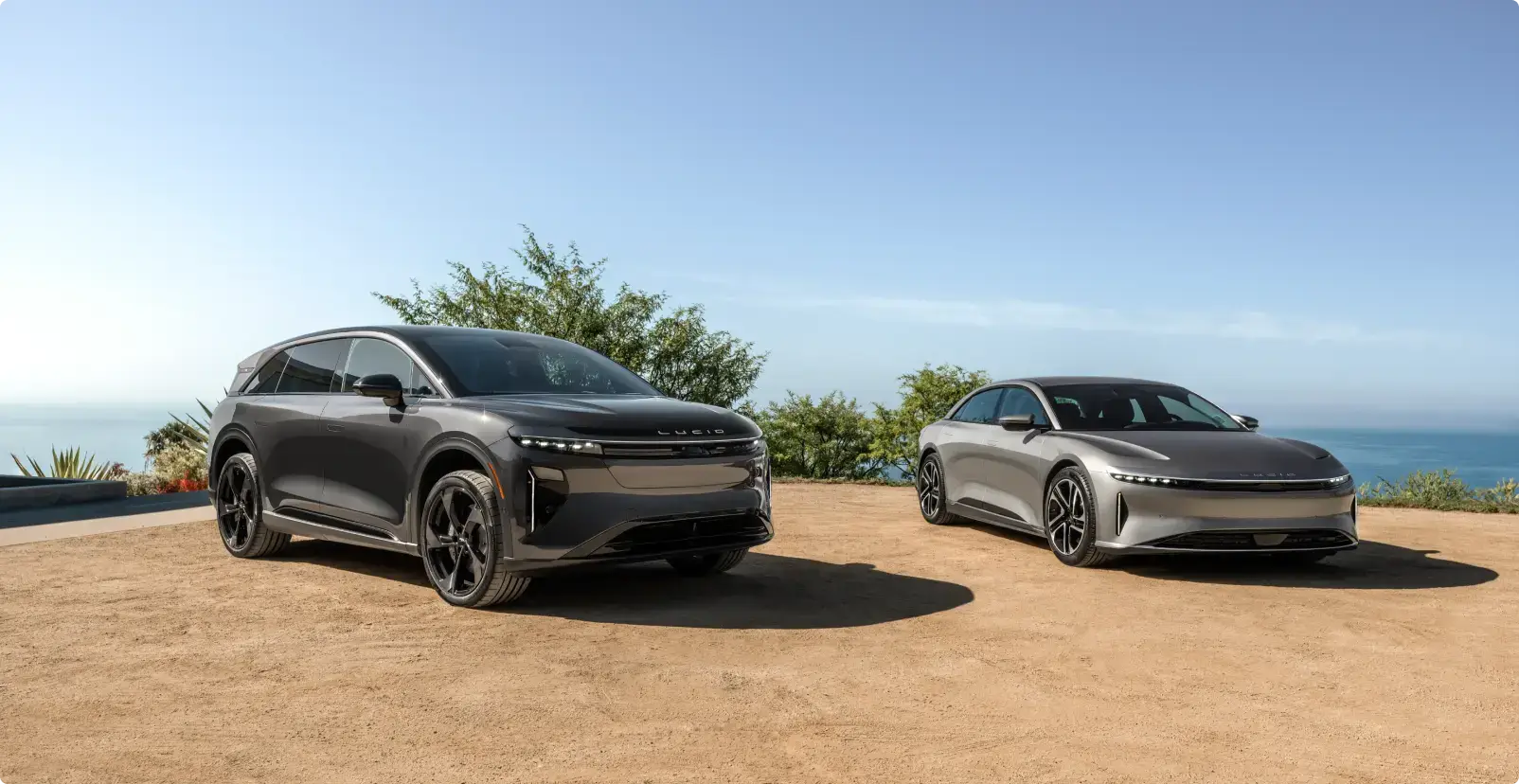 Two Lucid electric vehicles—a dark gray SUV and a silver sedan—parked outdoors with ocean and blue sky in the background.