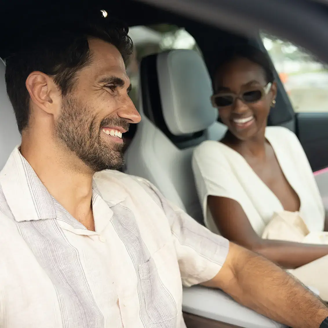 Two smiling people inside a car; a man in a white shirt in front and a woman in sunglasses and white outfit in the back.