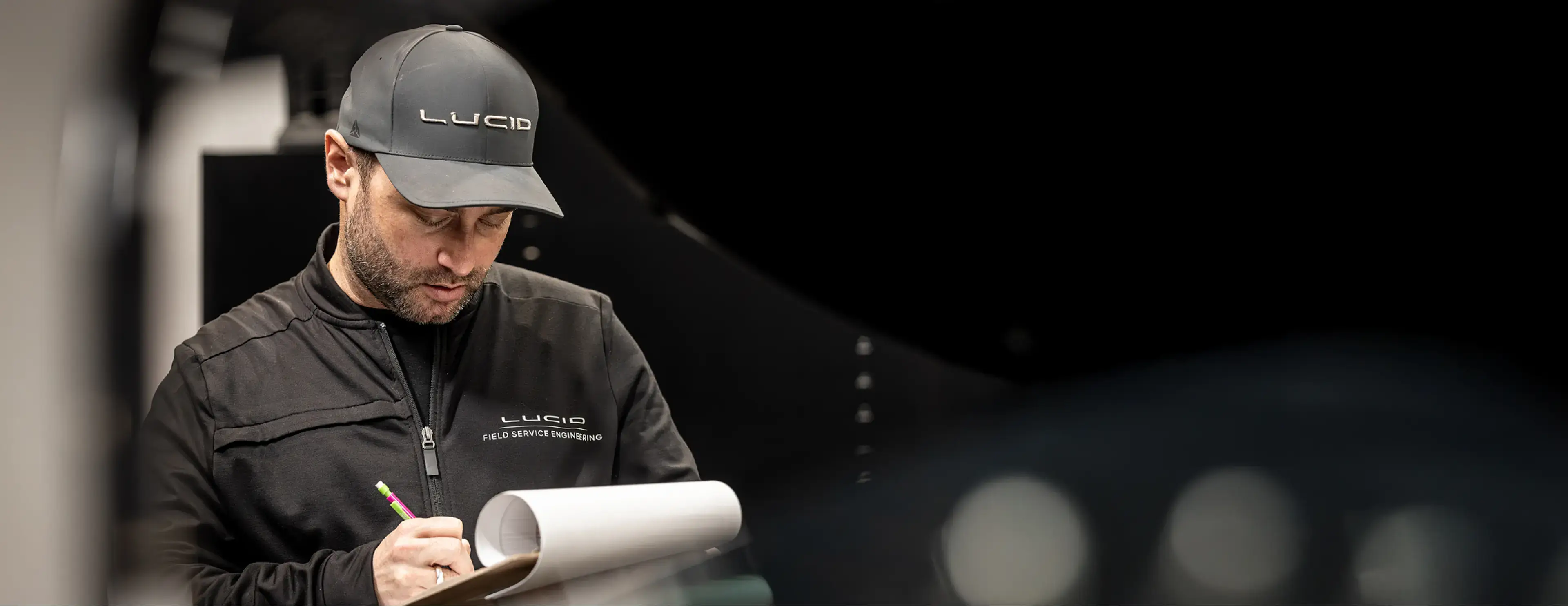 A Lucid Field Service Engineering technician in black uniform and gray cap taking notes on a clipboard in a dark workspace.