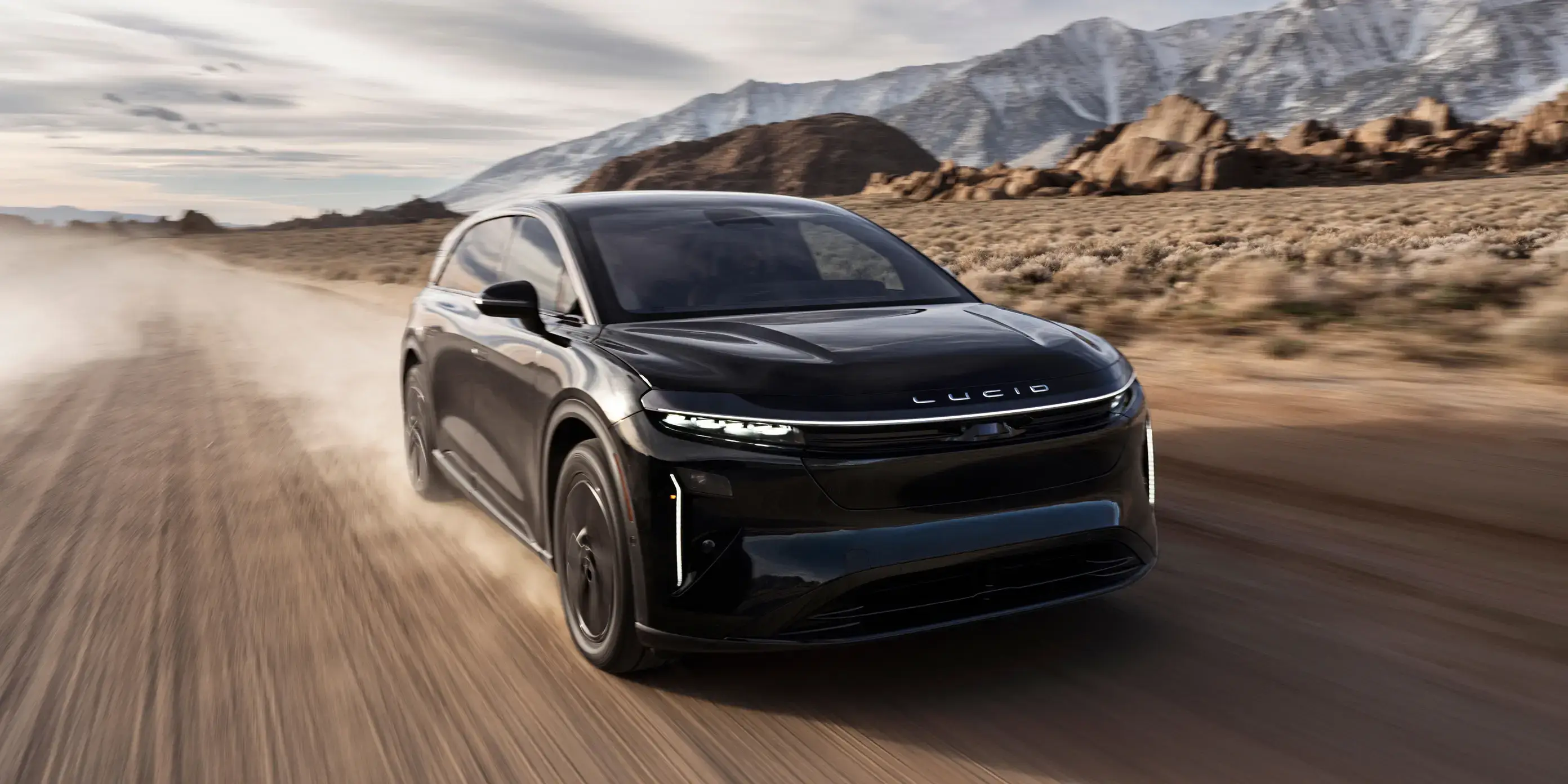 Lucid Gravity SUV driving on desert road with snow-capped mountains in background under cloudy sky.