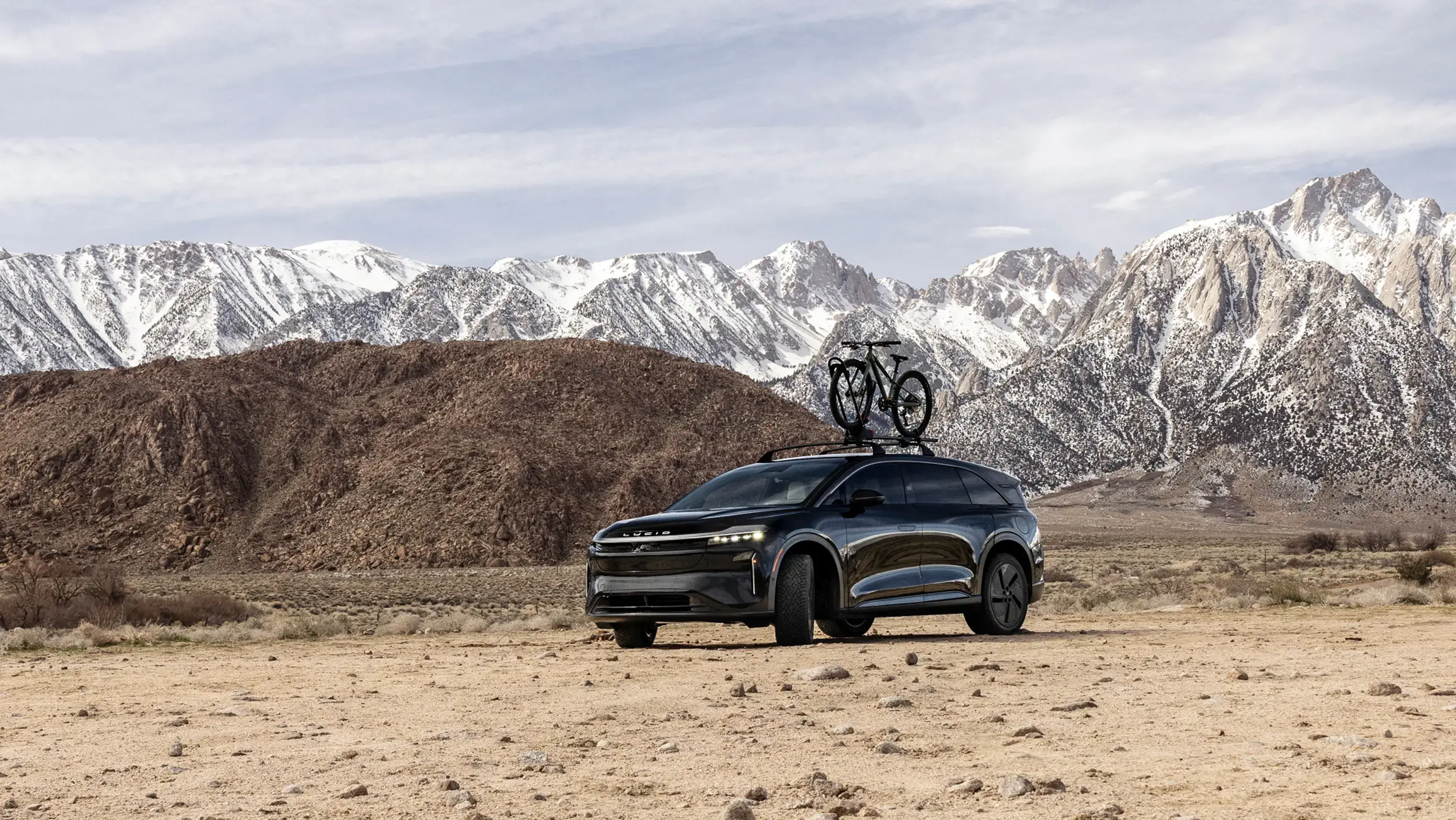Lucid Gravity SUV with a rooftop bike rack parked in a desert landscape with snow-capped mountains behind.