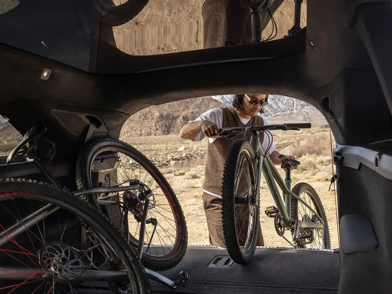 Person loading a mountain bike into the back of a Lucid Gravity SUV in a desert landscape, viewed from inside the cargo area.