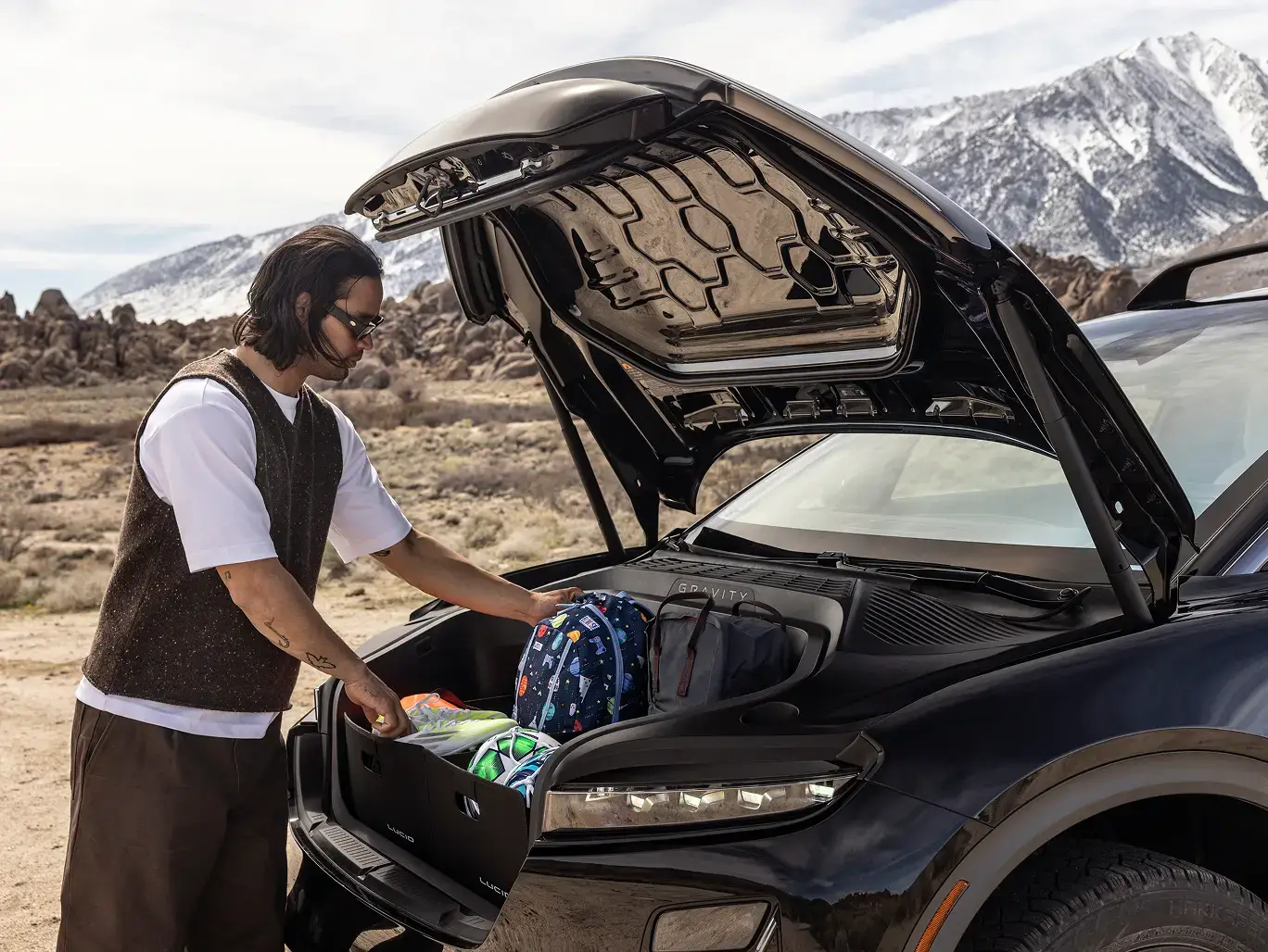A man loads luggage into the front trunk of a black electric SUV, with rocky desert terrain and snowy mountains behind him.