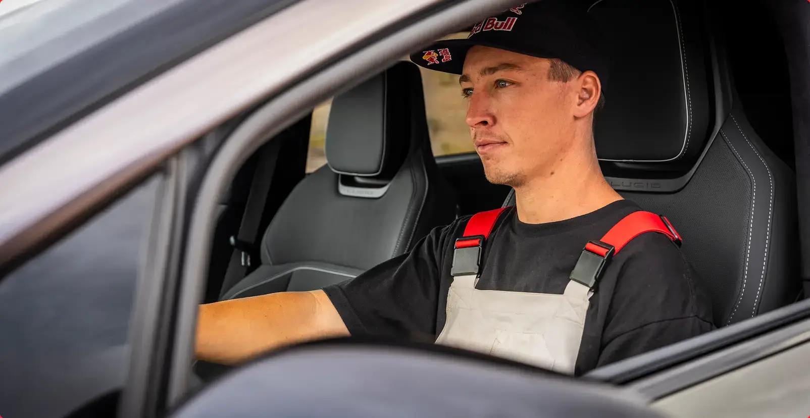 A young man wearing a Red Bull cap and racing harness sits in the driver's seat of a Lucid car, looking forward.