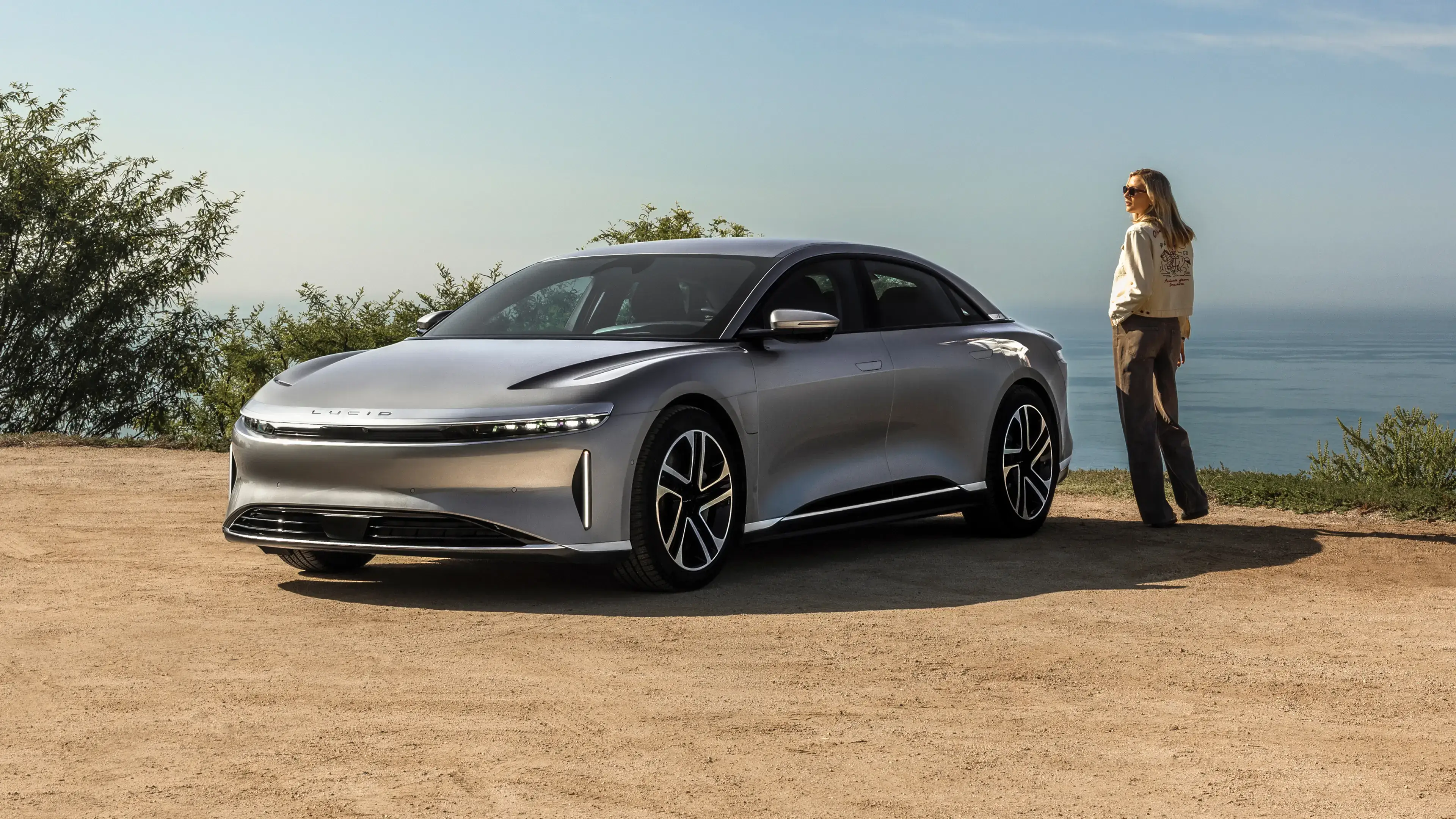 A silver Lucid Air electric sedan parked on a dirt overlook near the ocean, with a woman standing beside it.