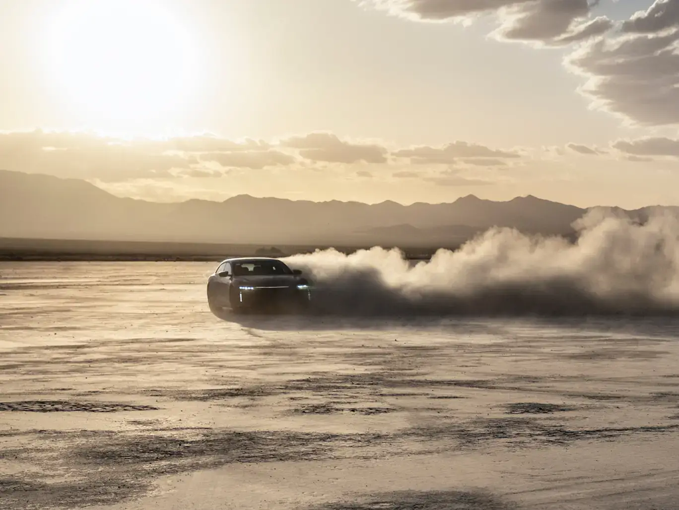 A dark sports car drifting on a desert flat, leaving a large smoke trail, with mountains and golden sunset sky.