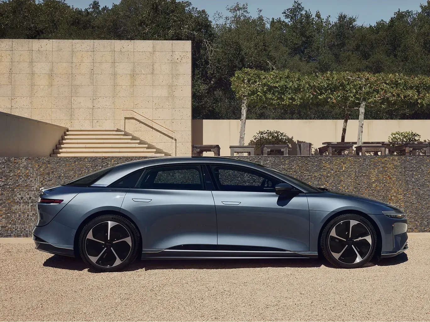 Fathom Blue Lucid Air sedan parked beside stone wall with modern architecture and manicured landscaping in background.