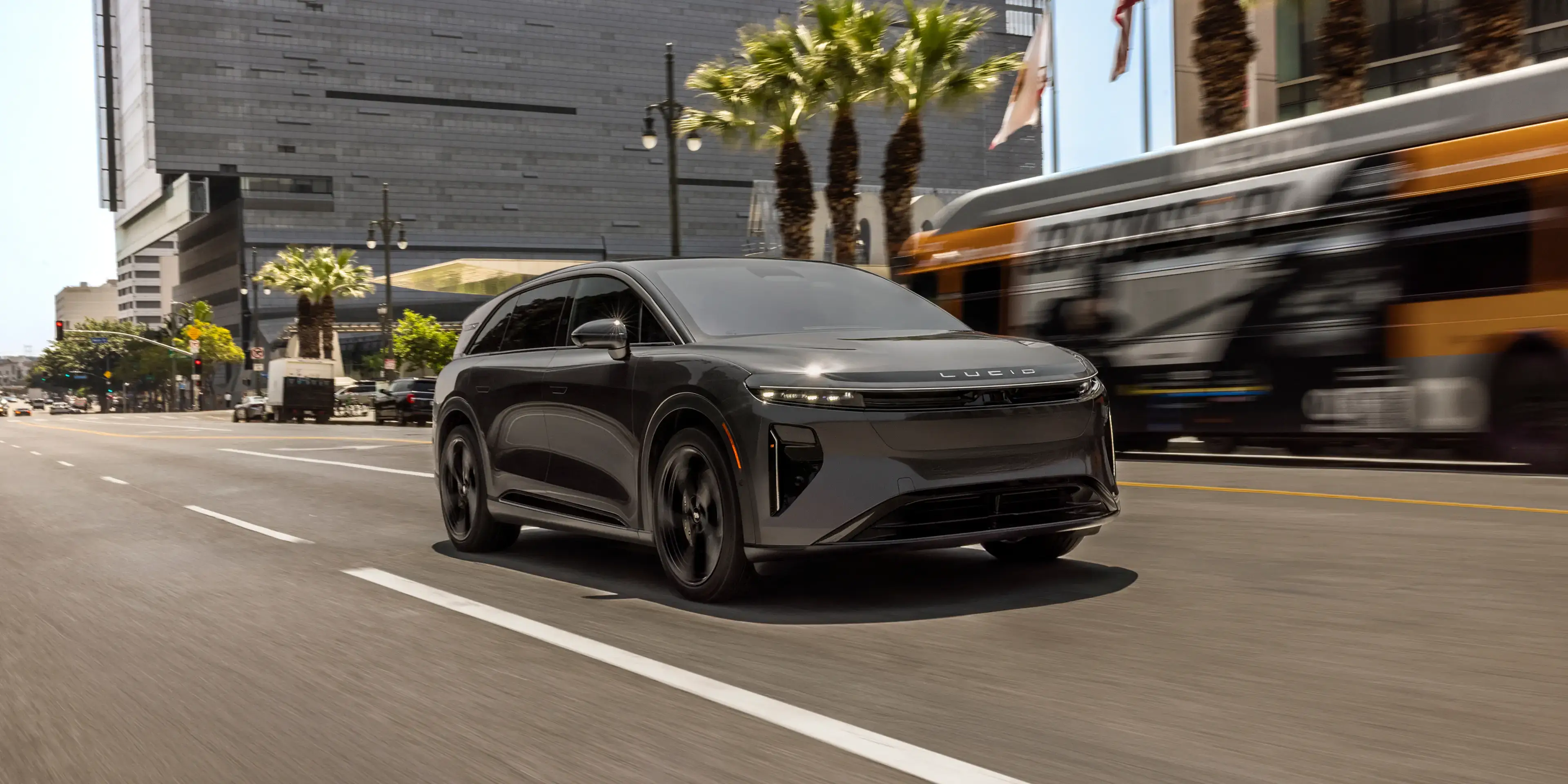 A dark gray Lucid electric SUV driving on a city street with palm trees and a blurred yellow bus in the background.