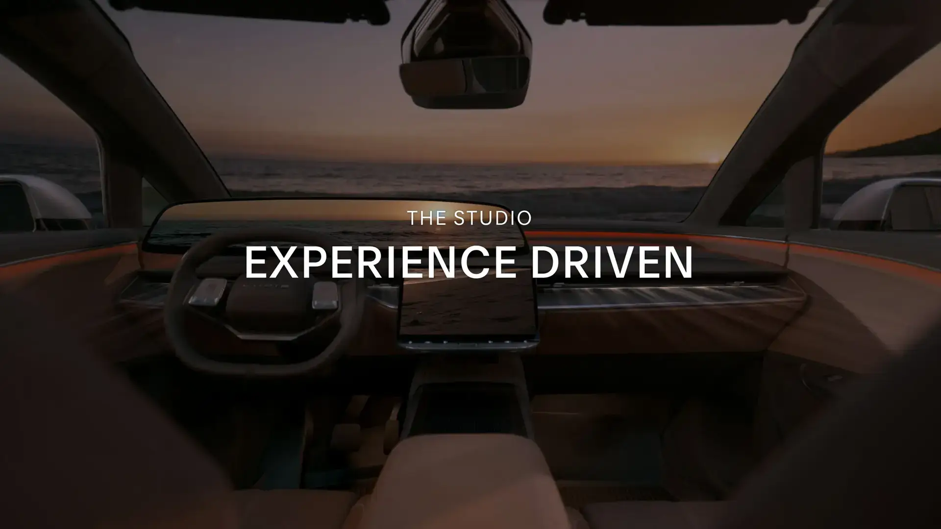 View from inside a luxury car interior facing an ocean sunset, with text overlay "THE STUDIO EXPERIENCE DRIVEN"