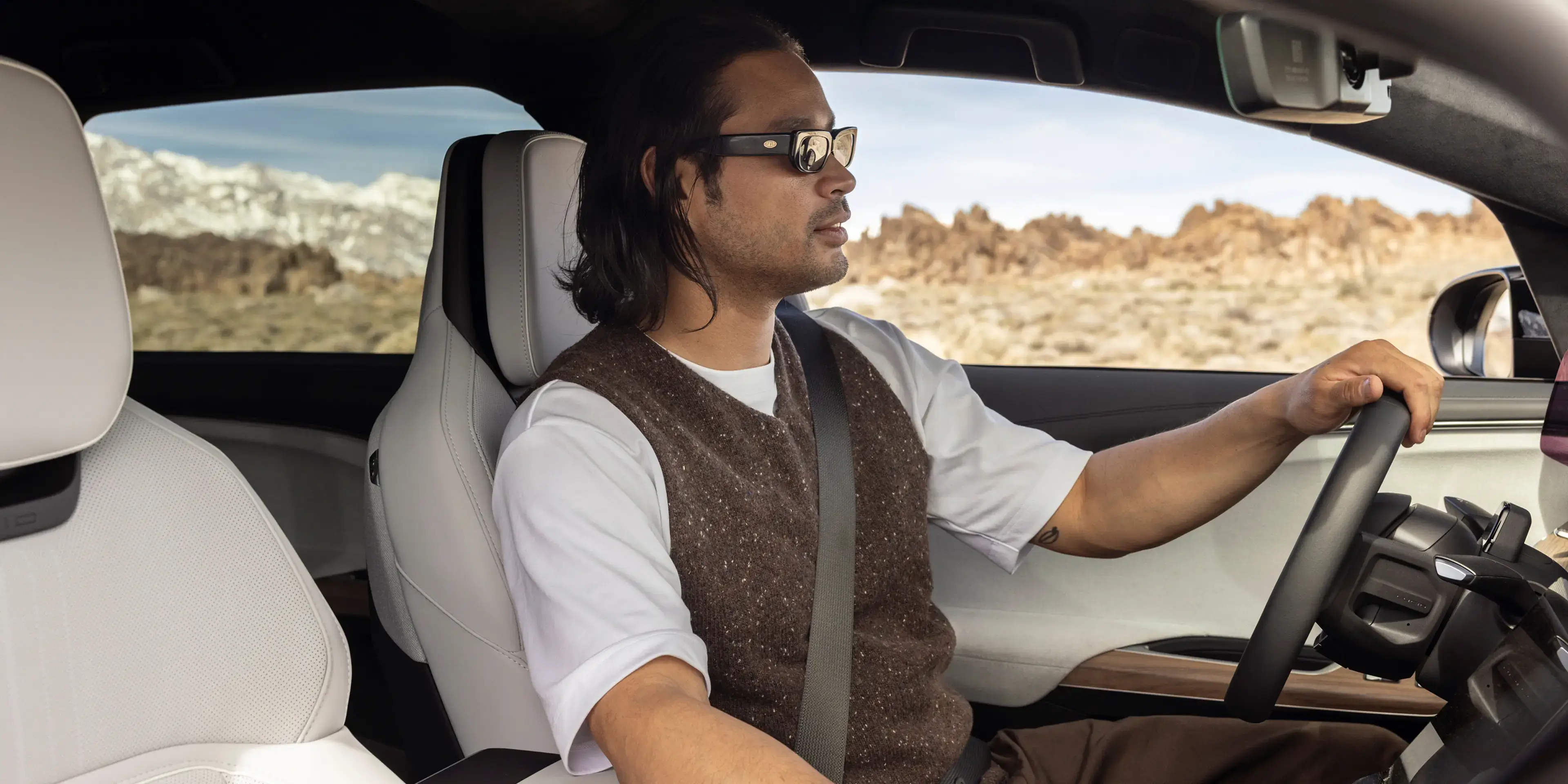 Person driving through desert landscape in luxury car interior, wearing brown vest and sunglasses with mountains visible outside.