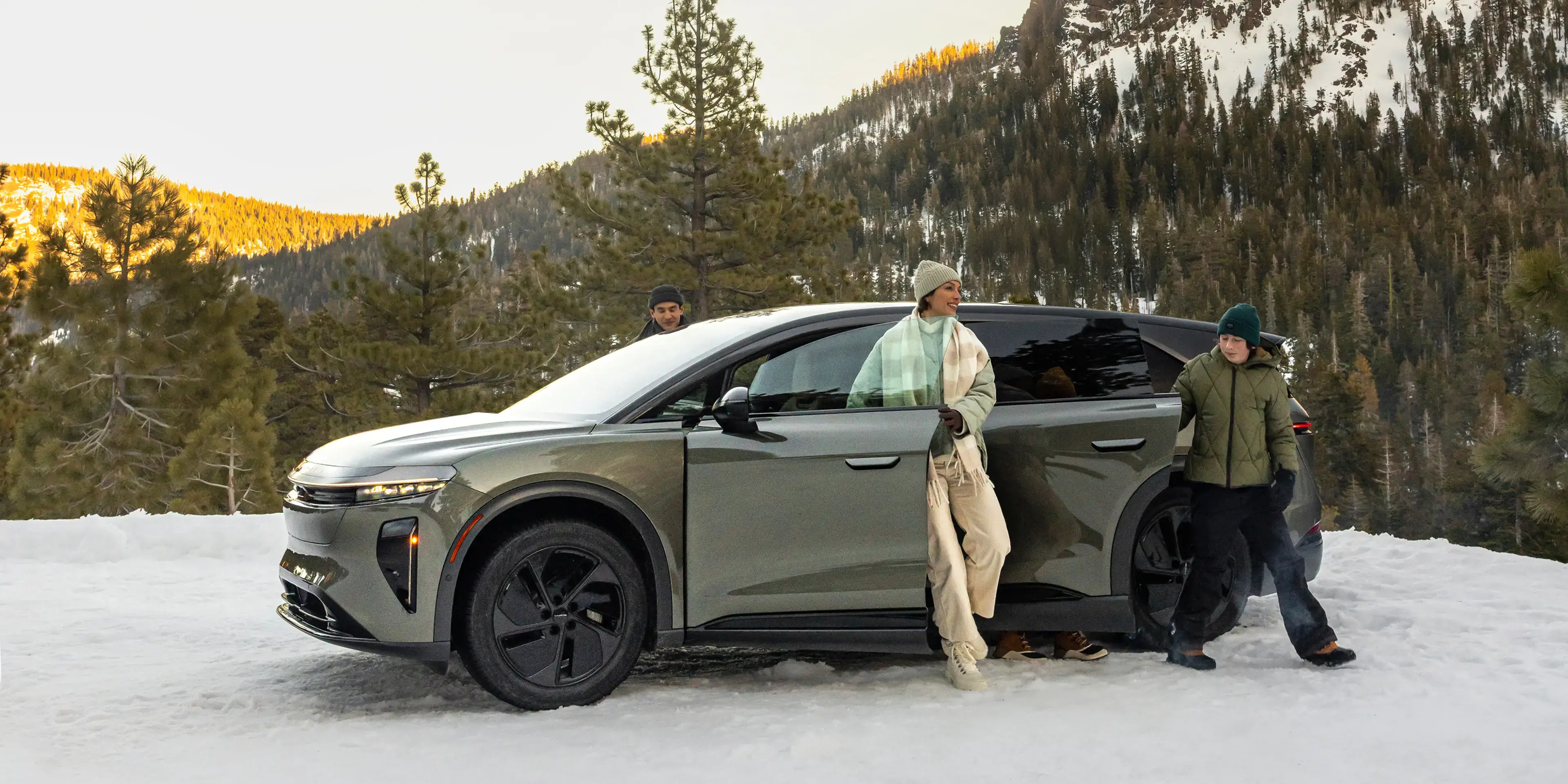 Lucid Gravity SUV parked in snowy mountain landscape with people in winter clothing against sunset-lit peaks.
