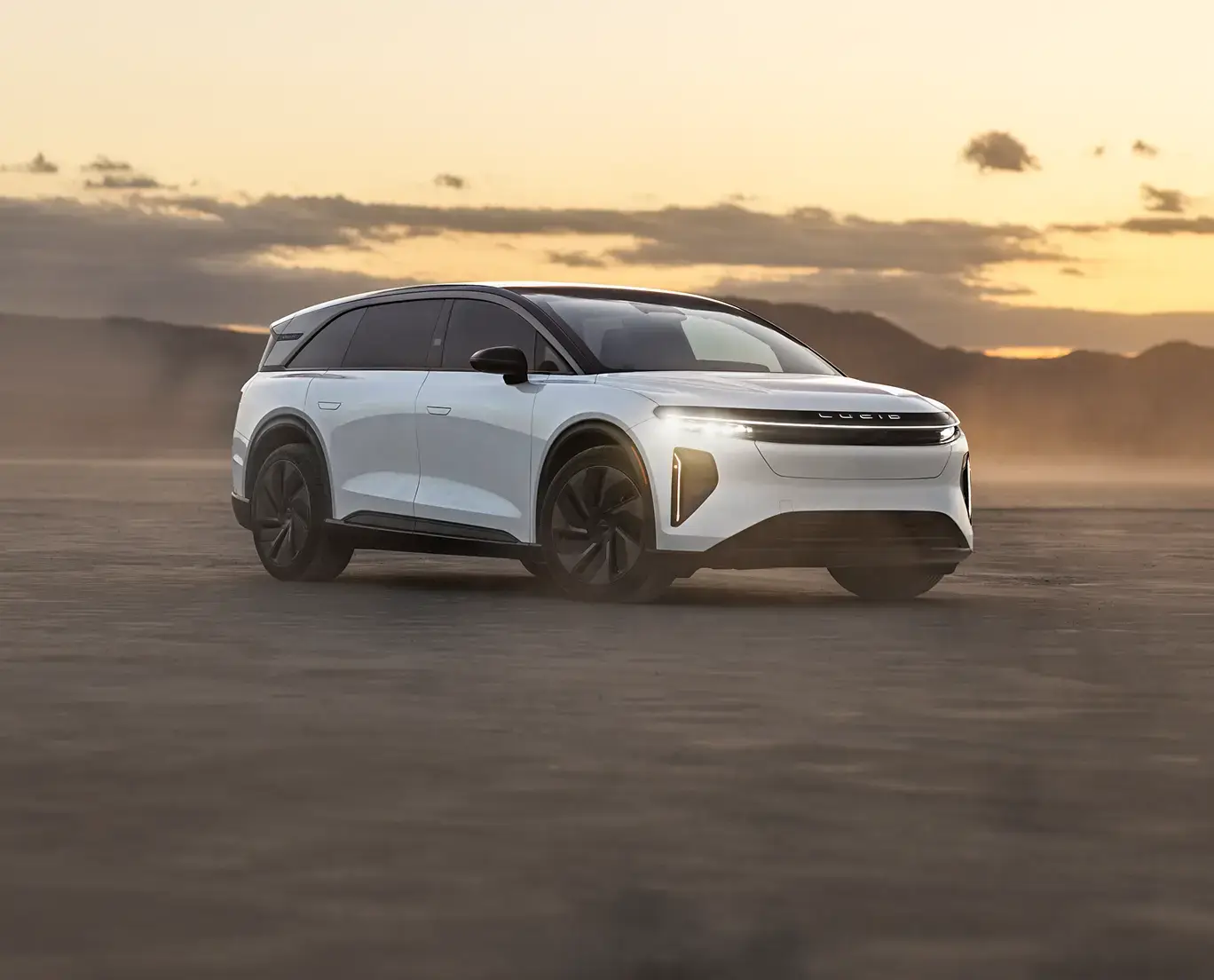 White Lucid electric SUV parked on a desert flat at sunset, with golden sky and misty mountains in the background.