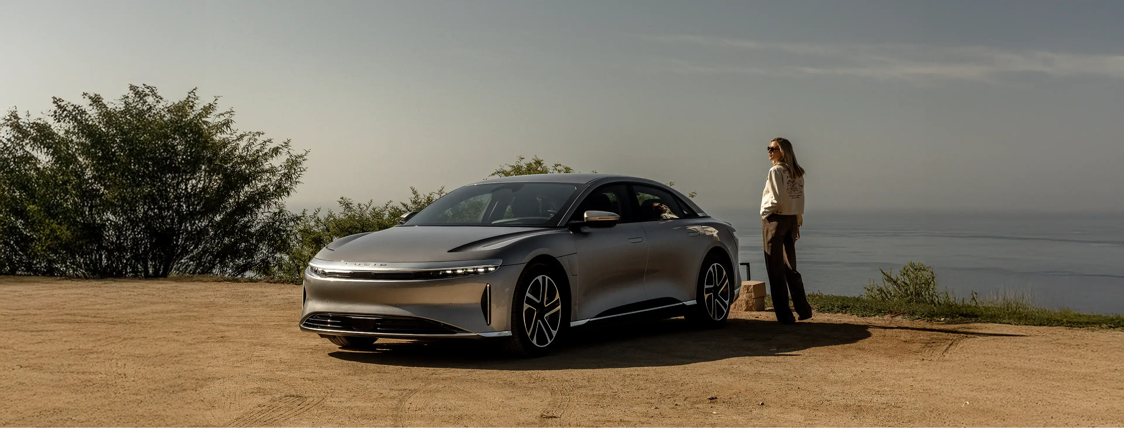A silver Lucid Air electric sedan parked on a dirt overlook near the ocean, with a woman standing beside it.