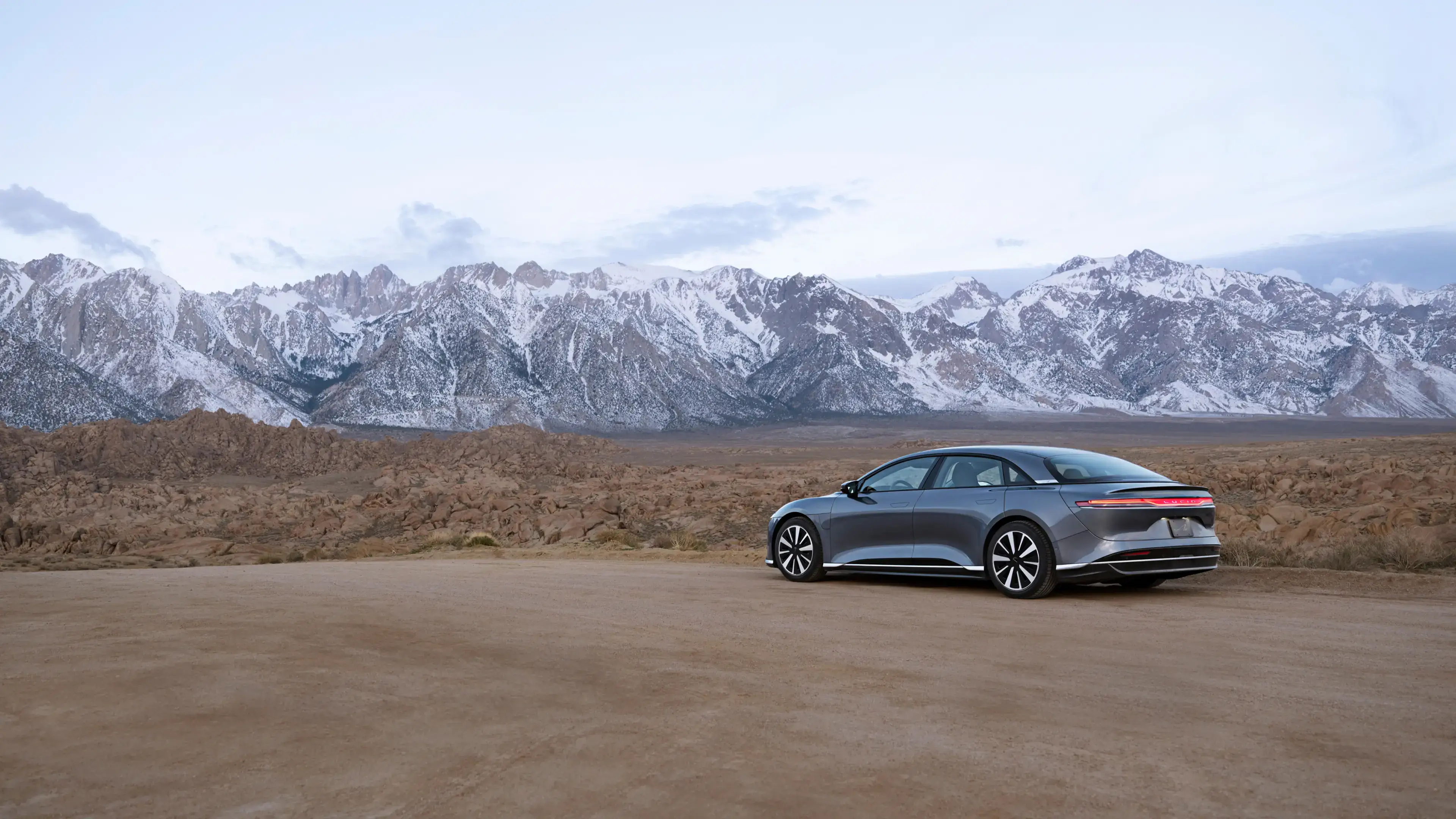 Lucid Air sedan parked on desert terrain with snow-capped mountain range in the background under clear sky.