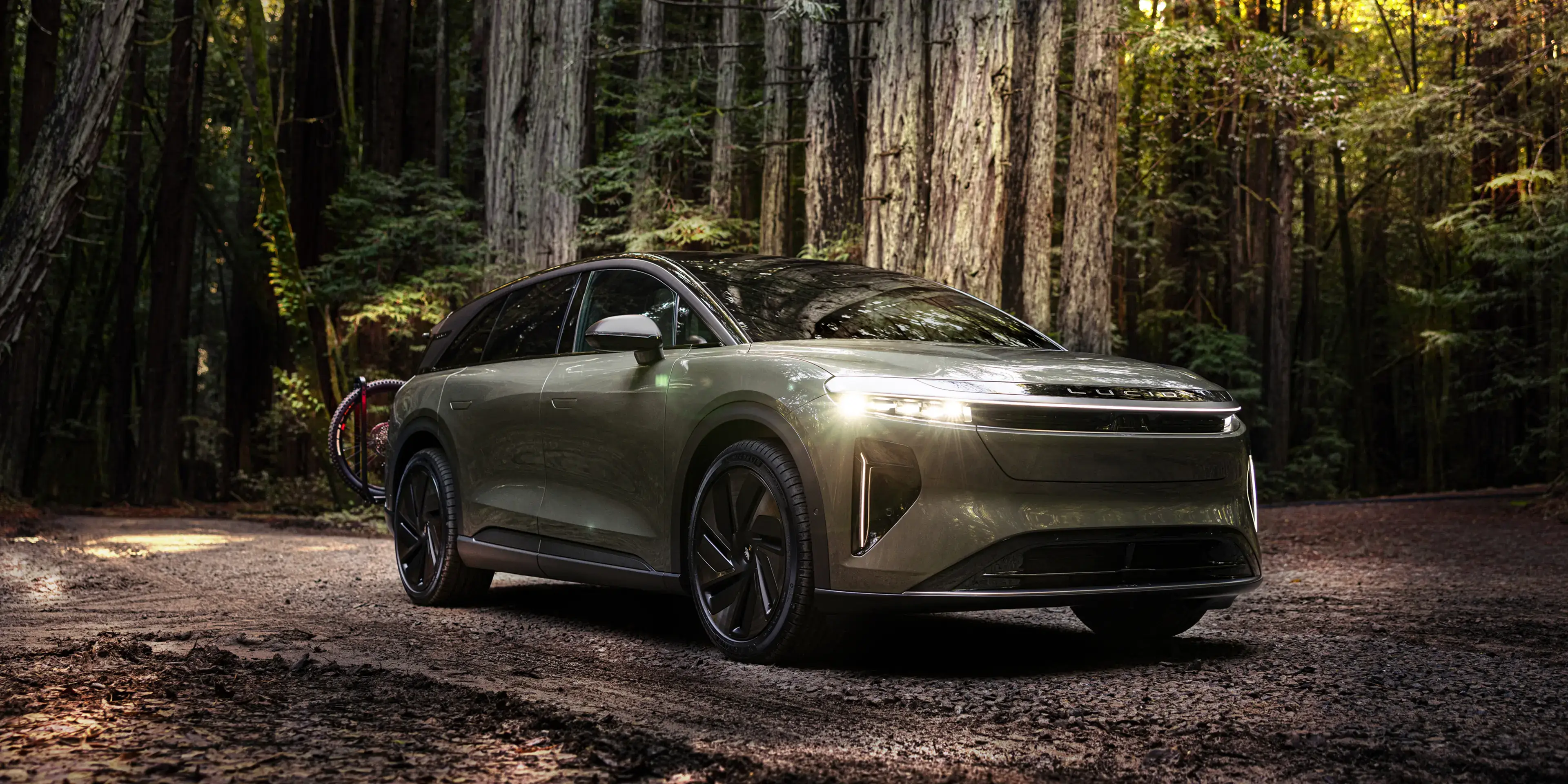 A Lucid Gravity SUV parked on a forest dirt path, headlights on, with a bicycle mounted on the rear.