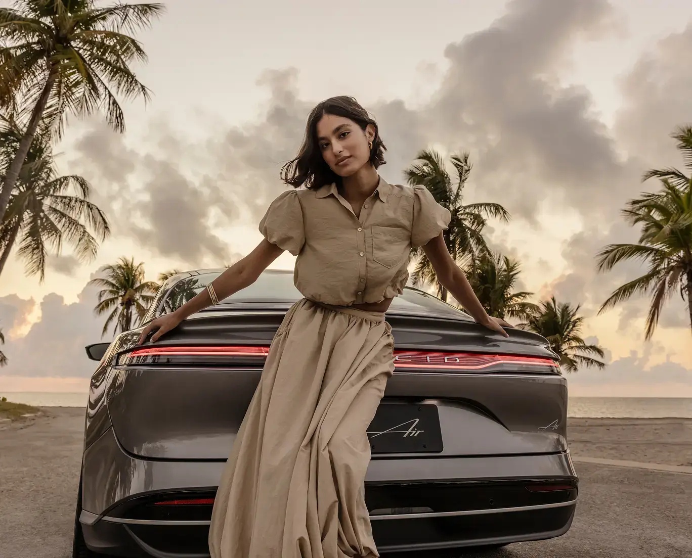 Woman in beige outfit leaning on the rear of a gray Lucid Air luxury EV near a beach with palm trees at dusk.