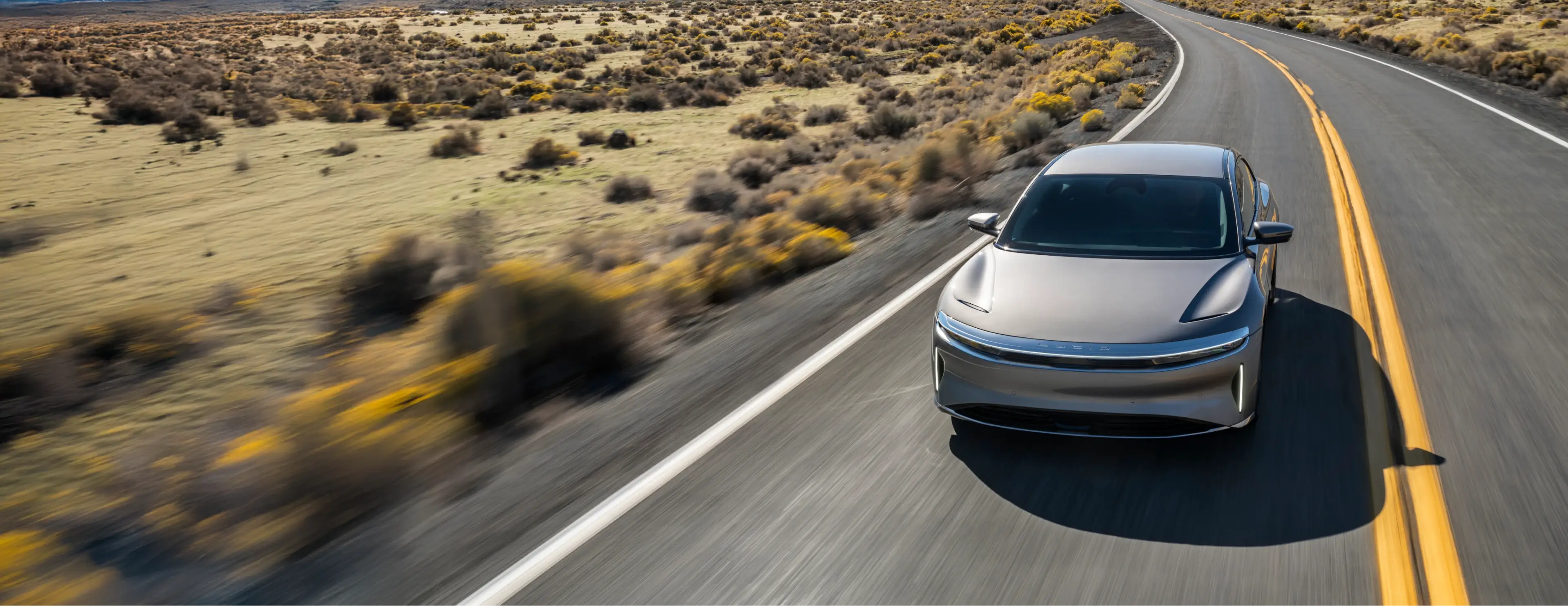 Silver electric vehicle driving on a winding desert highway with sparse vegetation and yellow wildflowers in the landscape.