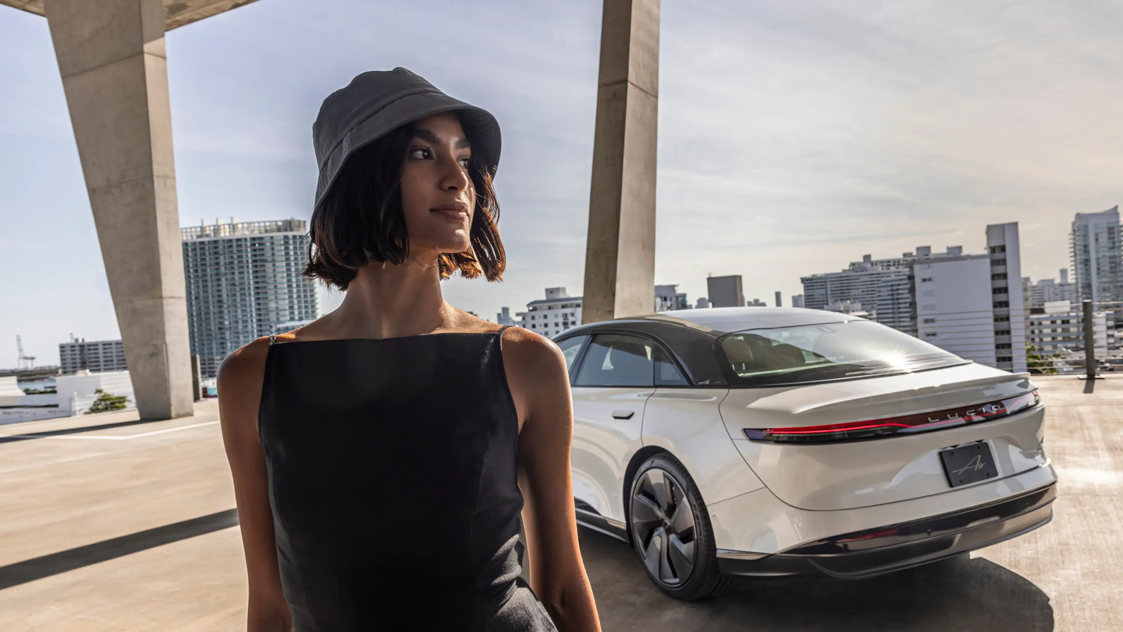 Person in black dress and bucket hat standing beside a white Lucid electric car with city skyline in background.