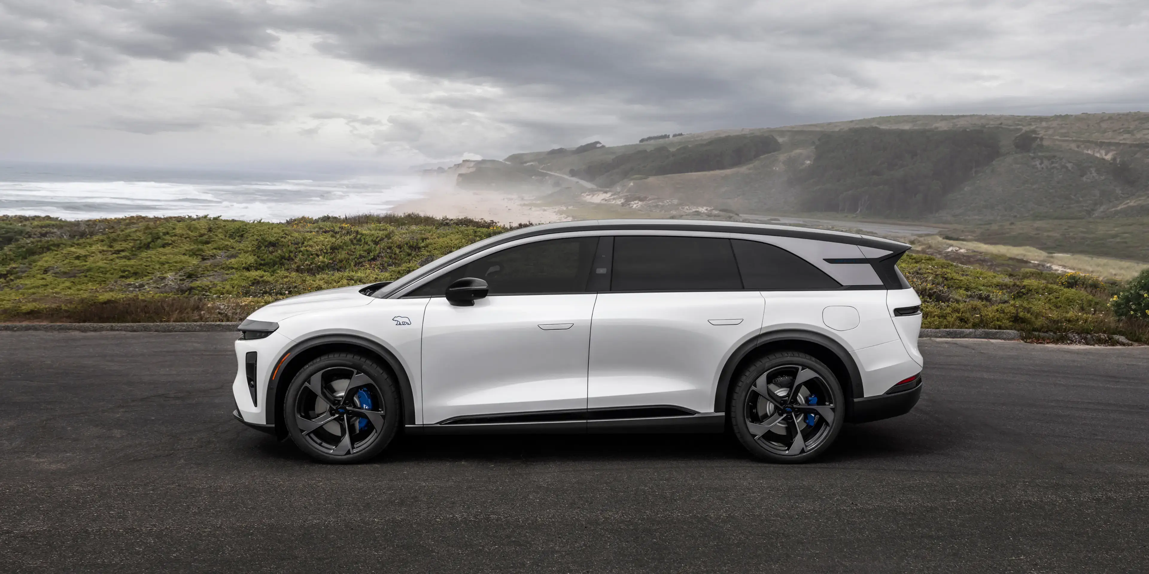 White electric SUV with black roof and blue brake calipers parked on a coastal overlook with misty cliffs and ocean behind.