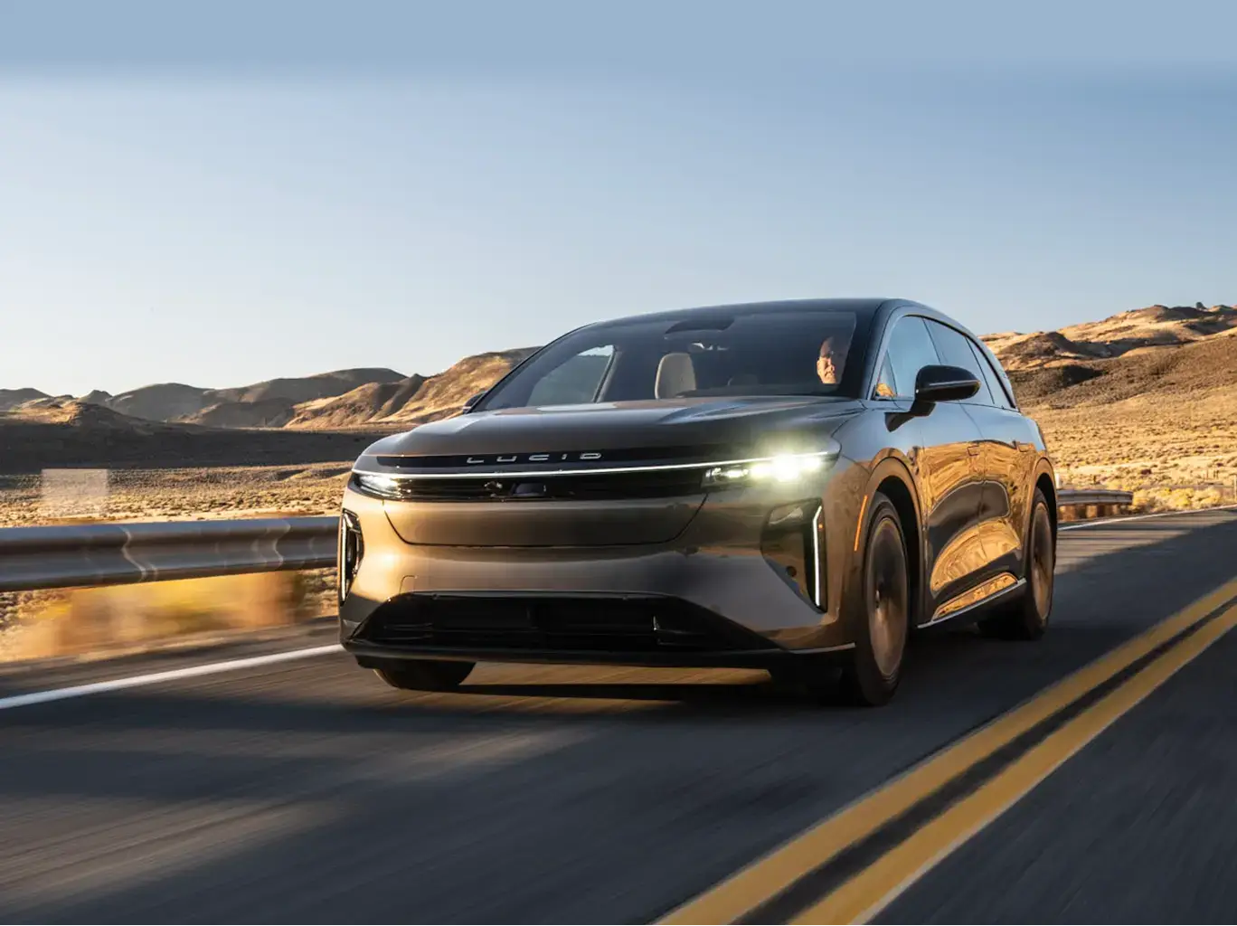 سيارة Lucid Gravity تسير على طريق صحراوي سريع مع تشغيل المصابيح الأمامية، وتتميز خلفية الصورة بالتلال ذهبية اللون تحت السماء الزرقاء.