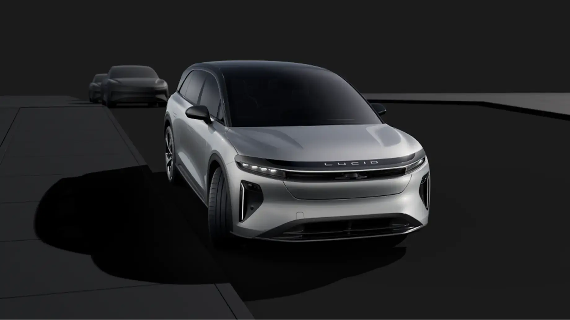 Silver Lucid Gravity electric SUV shown from the front against a dark background, with a second dark vehicle visible behind it.