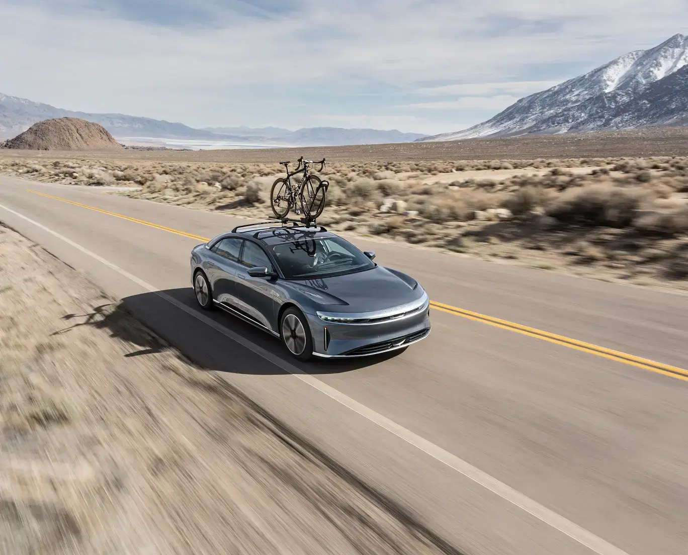 Gray electric sedan with bike on roof rack driving through desert landscape with snow-capped mountains in background.