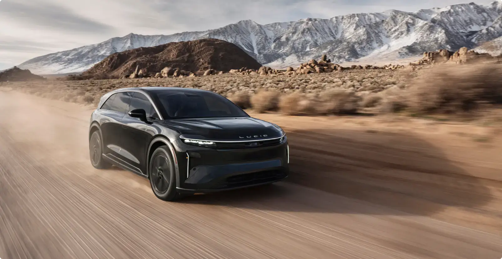 A Lucid Gravity SUV drives on a dusty desert road with snow-capped mountains in the background.