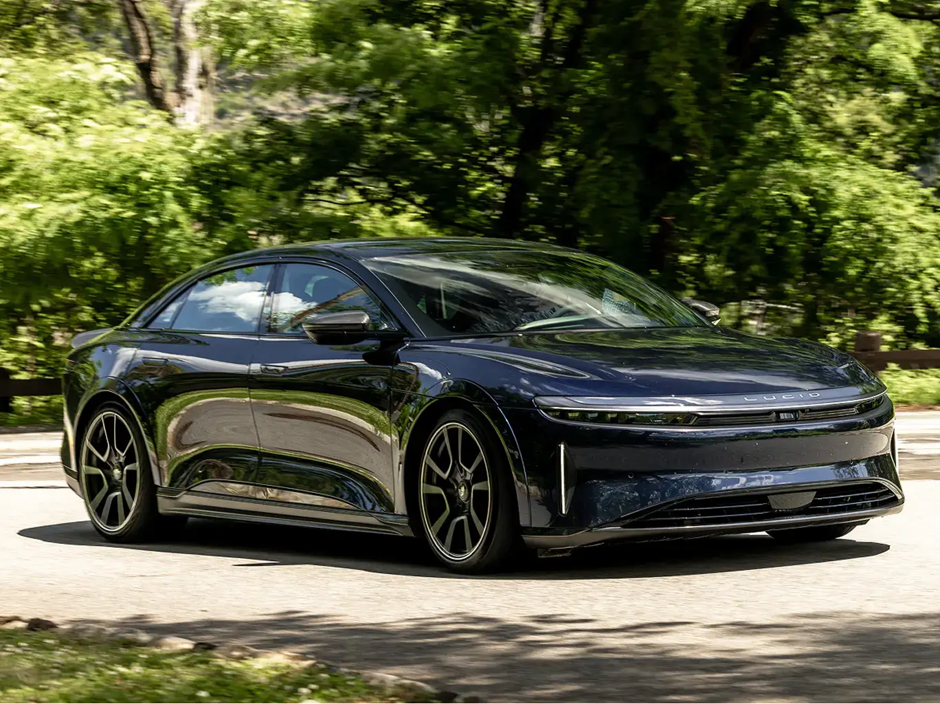A Lucid Air Sapphire electric sedan driving on a road surrounded by lush green trees.