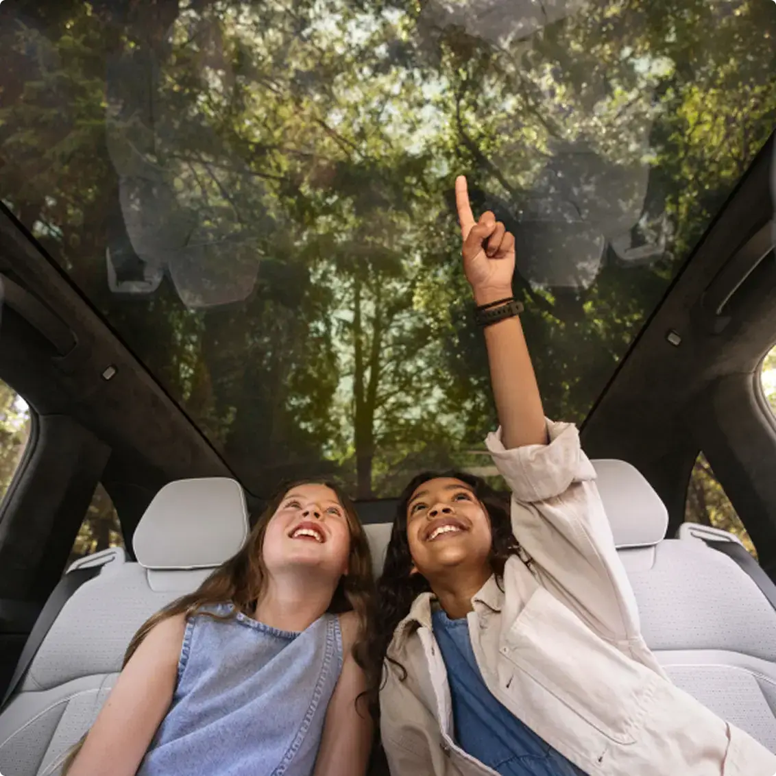 Two smiling girls sit in a car's back seat, looking up through a large panoramic sunroof at green forest trees.