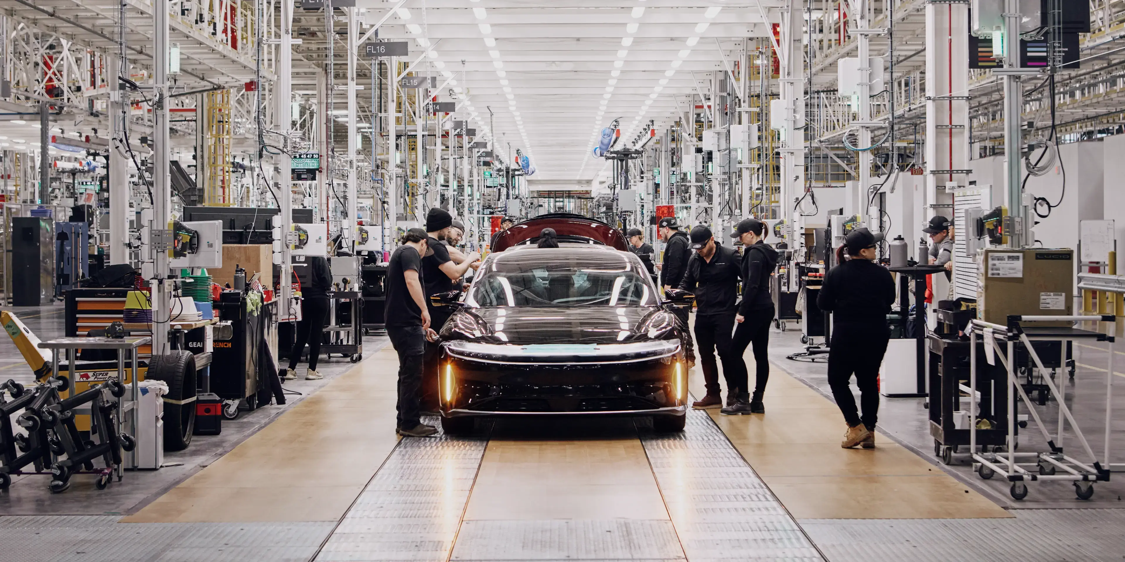 Workers in black uniforms assemble a dark red electric car on a bright, modern automotive factory floor.