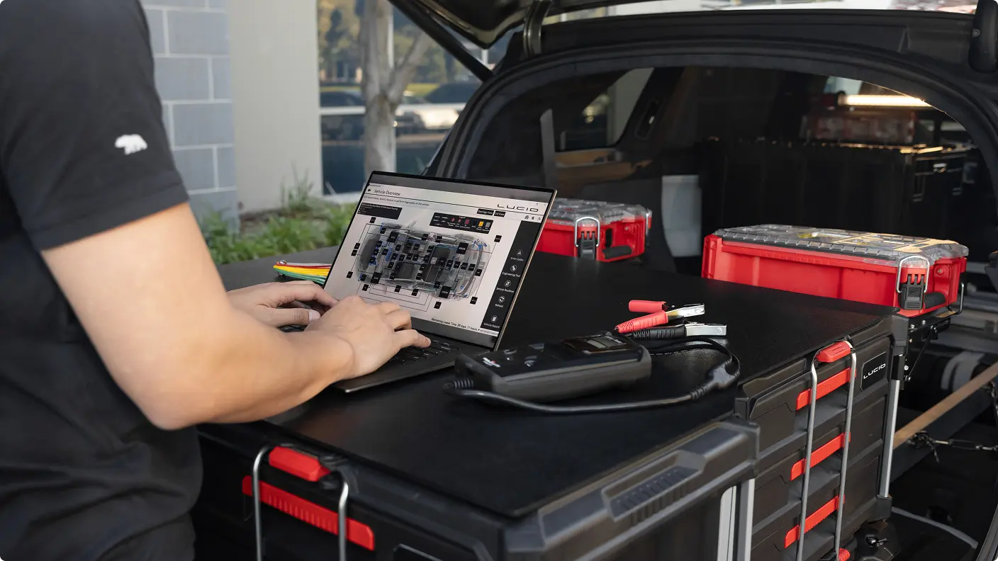 Person using Lucid laptop with vehicle diagnostic software at car trunk, surrounded by red tool cases and equipment.