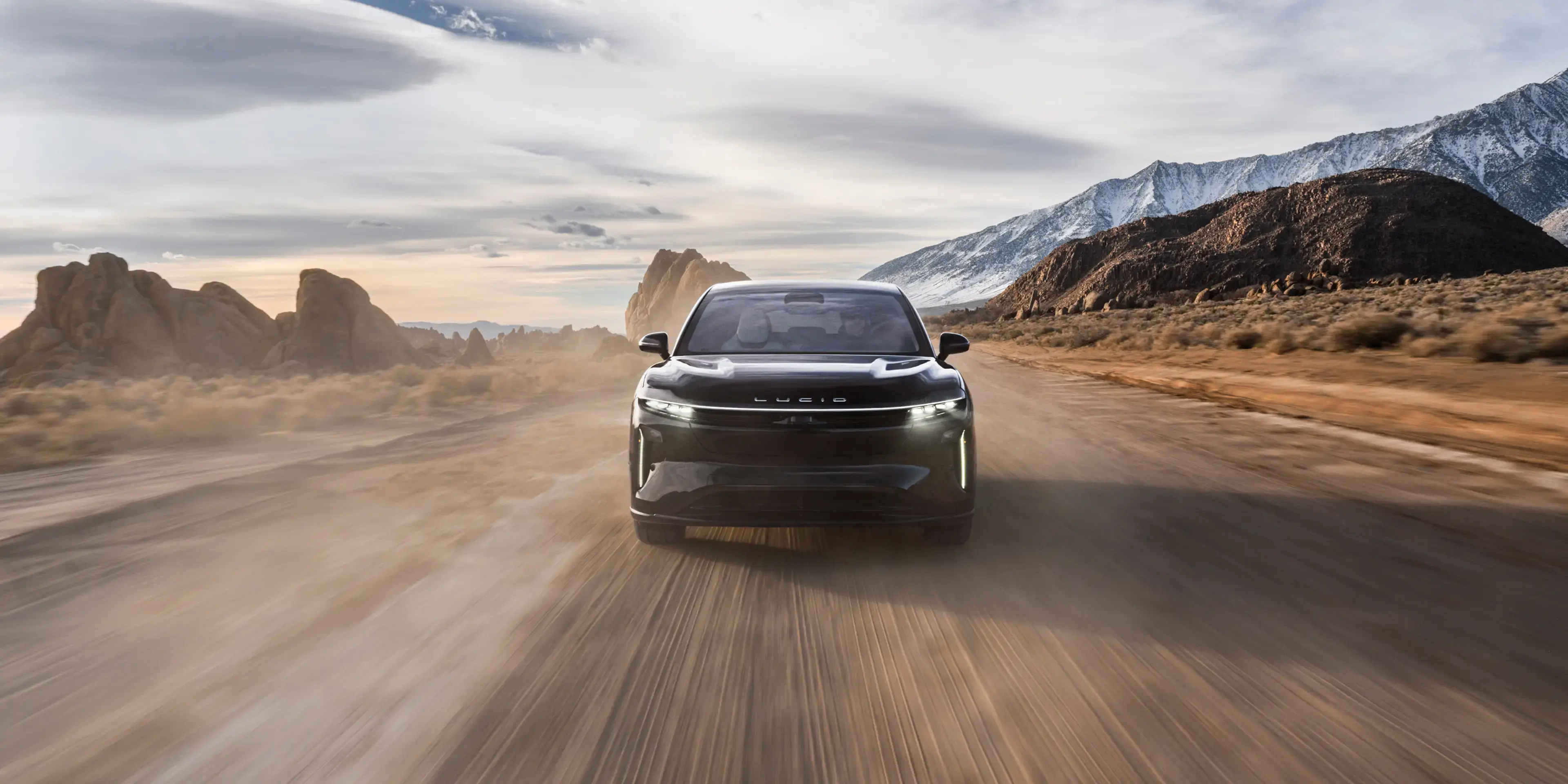 Lucid Gravity driving on desert road with snow-capped mountains and dramatic cloudy sky in background.