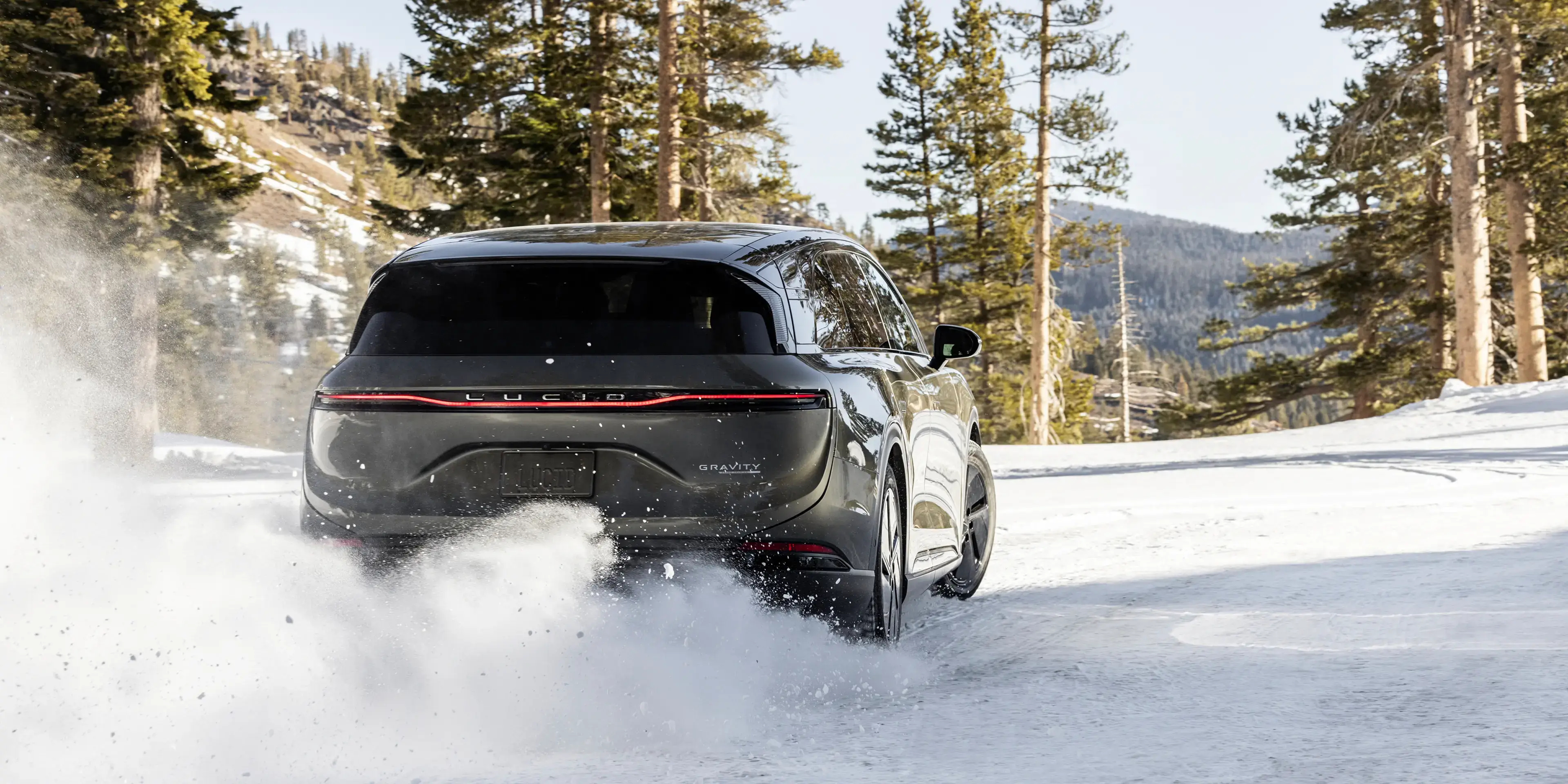 Lucid Gravity SUV driving through snow-covered mountain terrain with pine trees in the background.