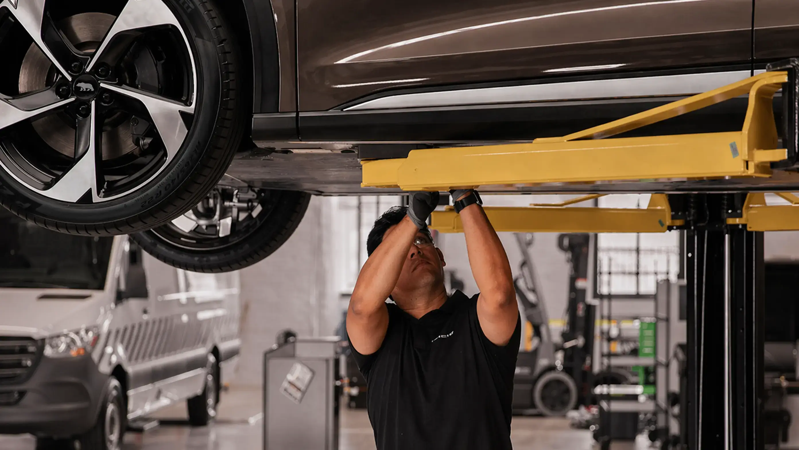 A mechanic in a black shirt inspects the underside of a car raised on a yellow lift in an auto repair shop.