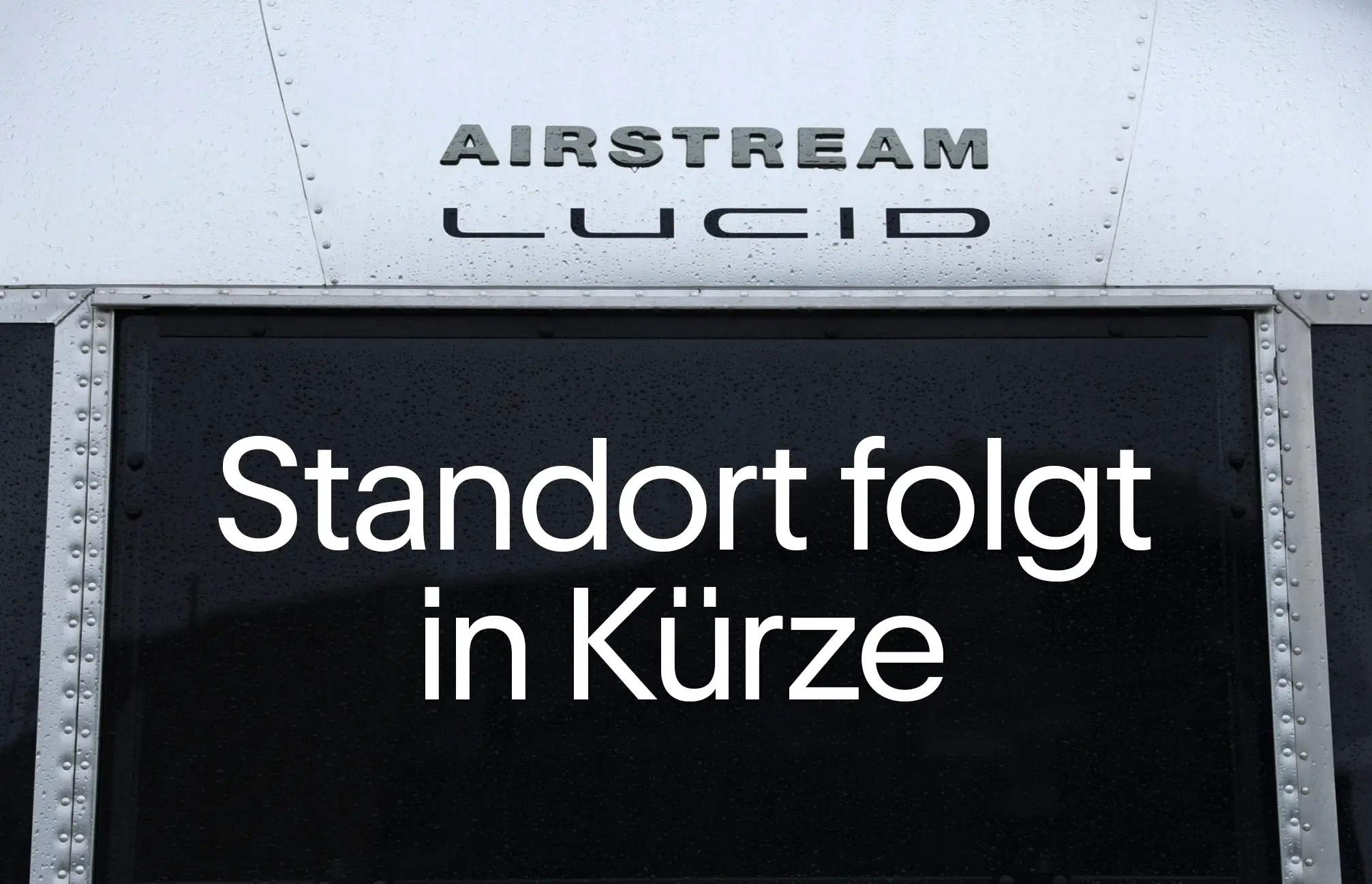 Nahaufnahme einer metallischen Airstream‑ähnlichen Oberfläche mit den erhabenen Schriftzügen „AIRSTREAM“ und „LUCID“ sowie „Standort folgt in Kürze“ auf dem dunklen Panel darunter.