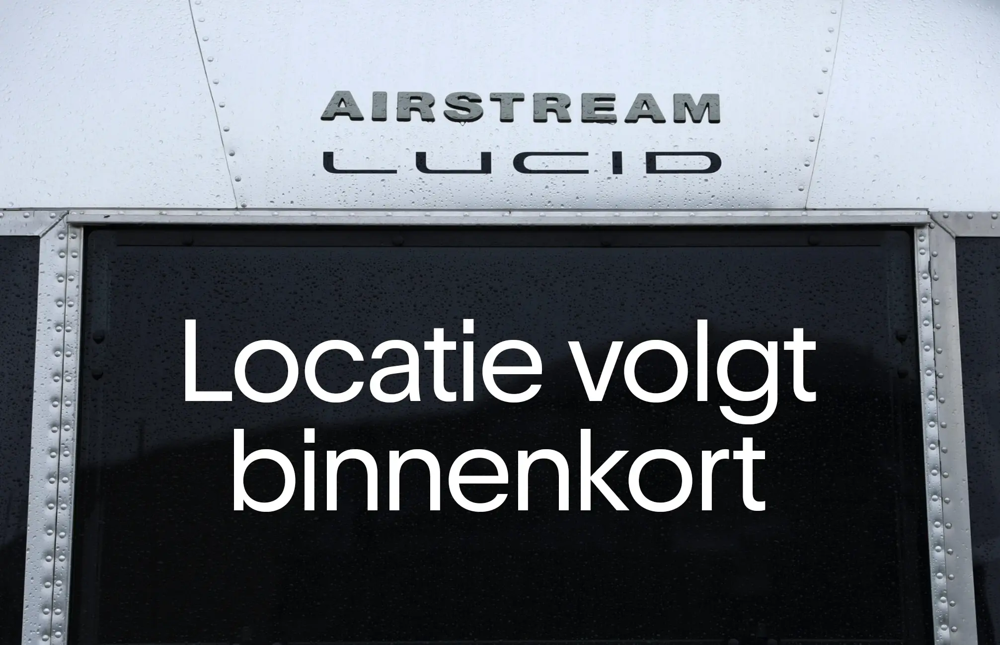 Close‑up van metalen Airstream‑achtige panelen met de verhoogde letters “AIRSTREAM” en “LUCID”, en “Locatie volgt binnenkort” op het donkere paneel eronder.