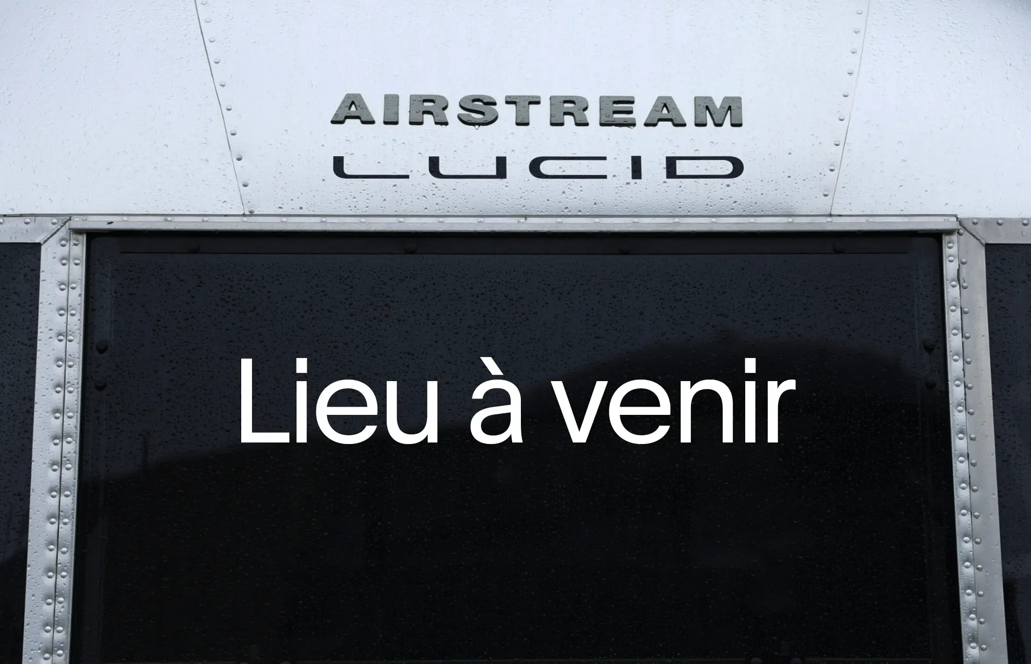 Gros plan sur un panneau métallique de style Airstream avec les inscriptions en relief « AIRSTREAM » et « LUCID », et « Lieu à venir » affiché sur le panneau sombre en dessous.