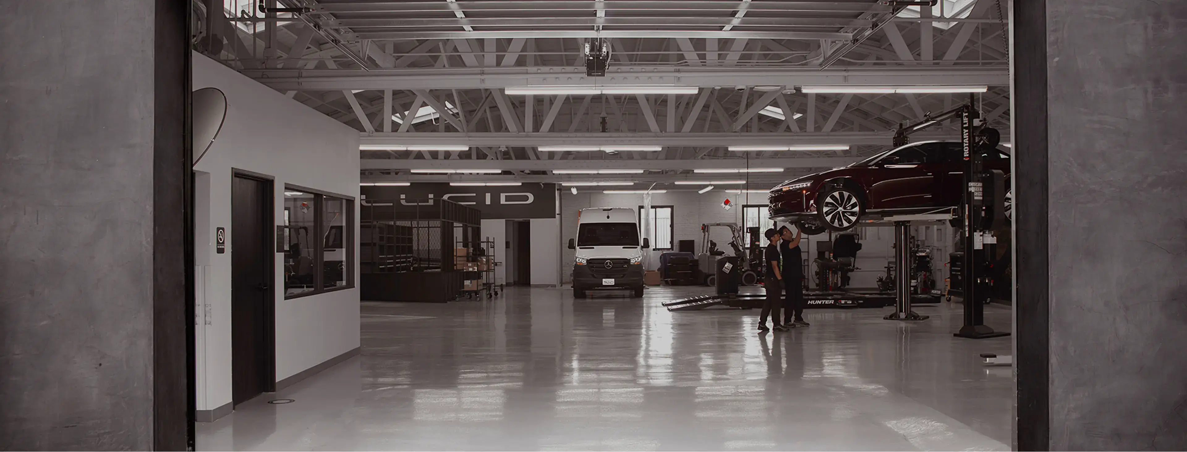 Lucid Motors service center with technicians inspecting a raised dark red car, a white van parked inside, and the Lucid logo on the wall.
