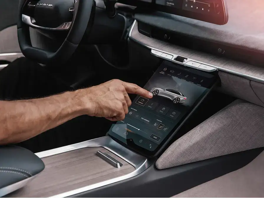 A hand tapping a large center touchscreen in a Lucid car interior, displaying vehicle controls and a car graphic.