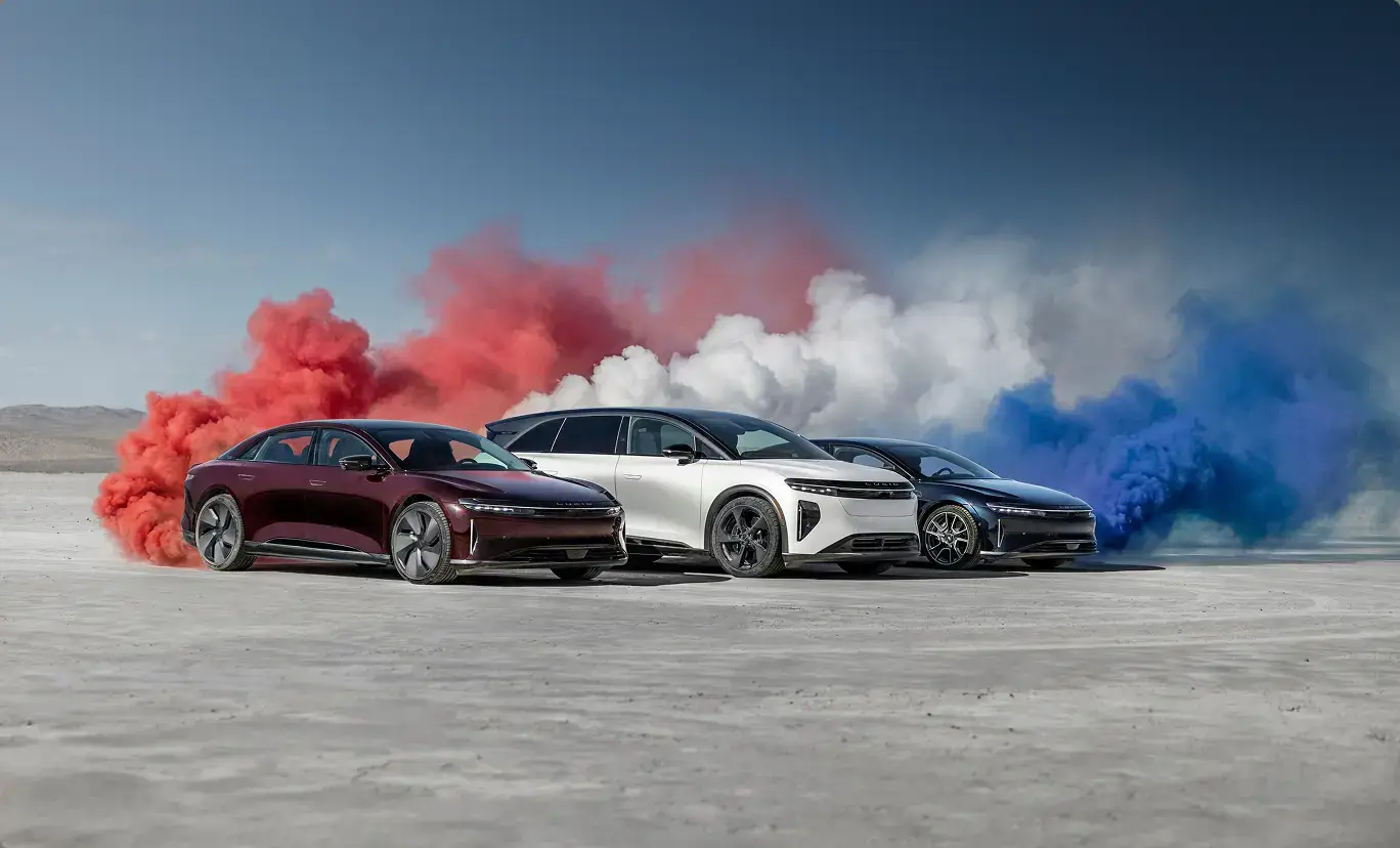 Three Lucid Air electric sedans in red, white, and blue on a salt flat with matching colored smoke clouds behind them.