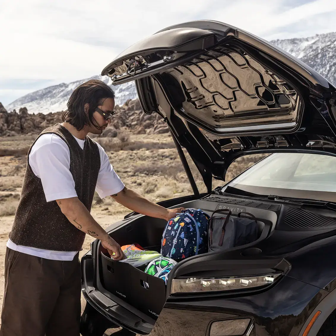 A man loads bags into the frunk of a black Lucid Gravity SUV in a desert landscape with snowy mountains behind.