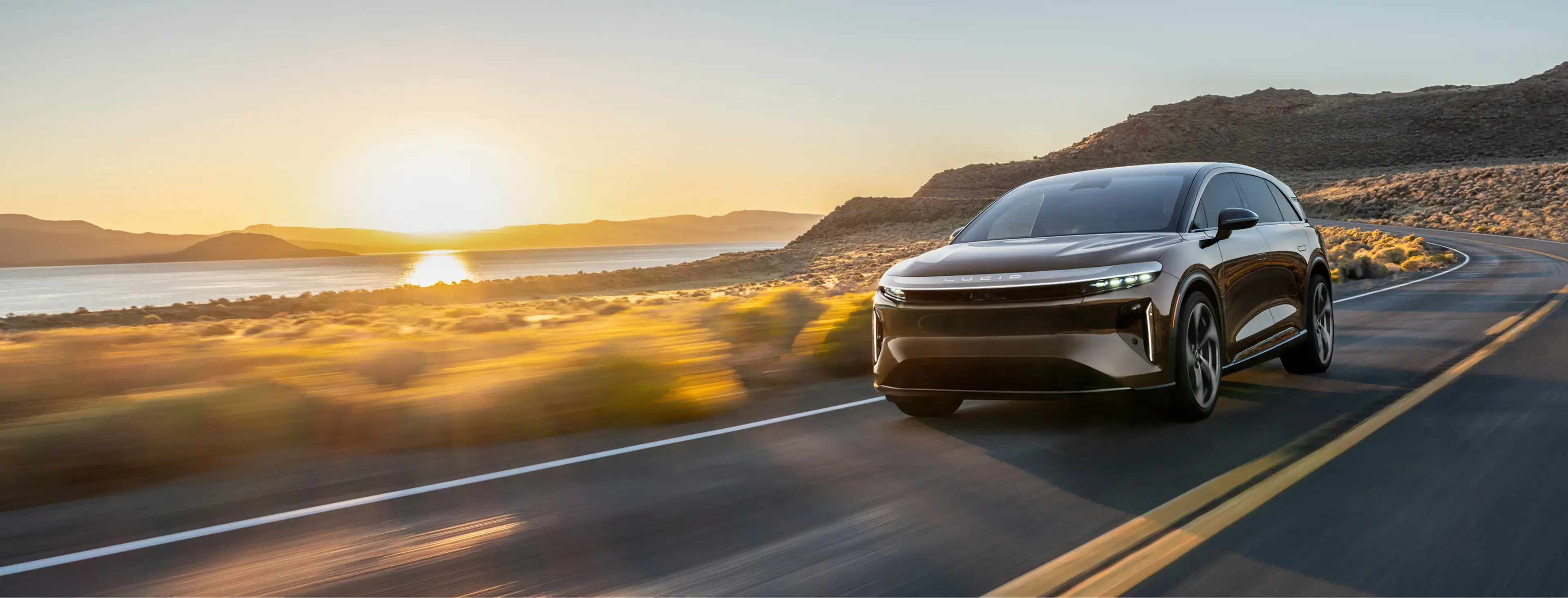 Sleek electric SUV driving on coastal highway at sunset, with golden light reflecting off mountains and water.