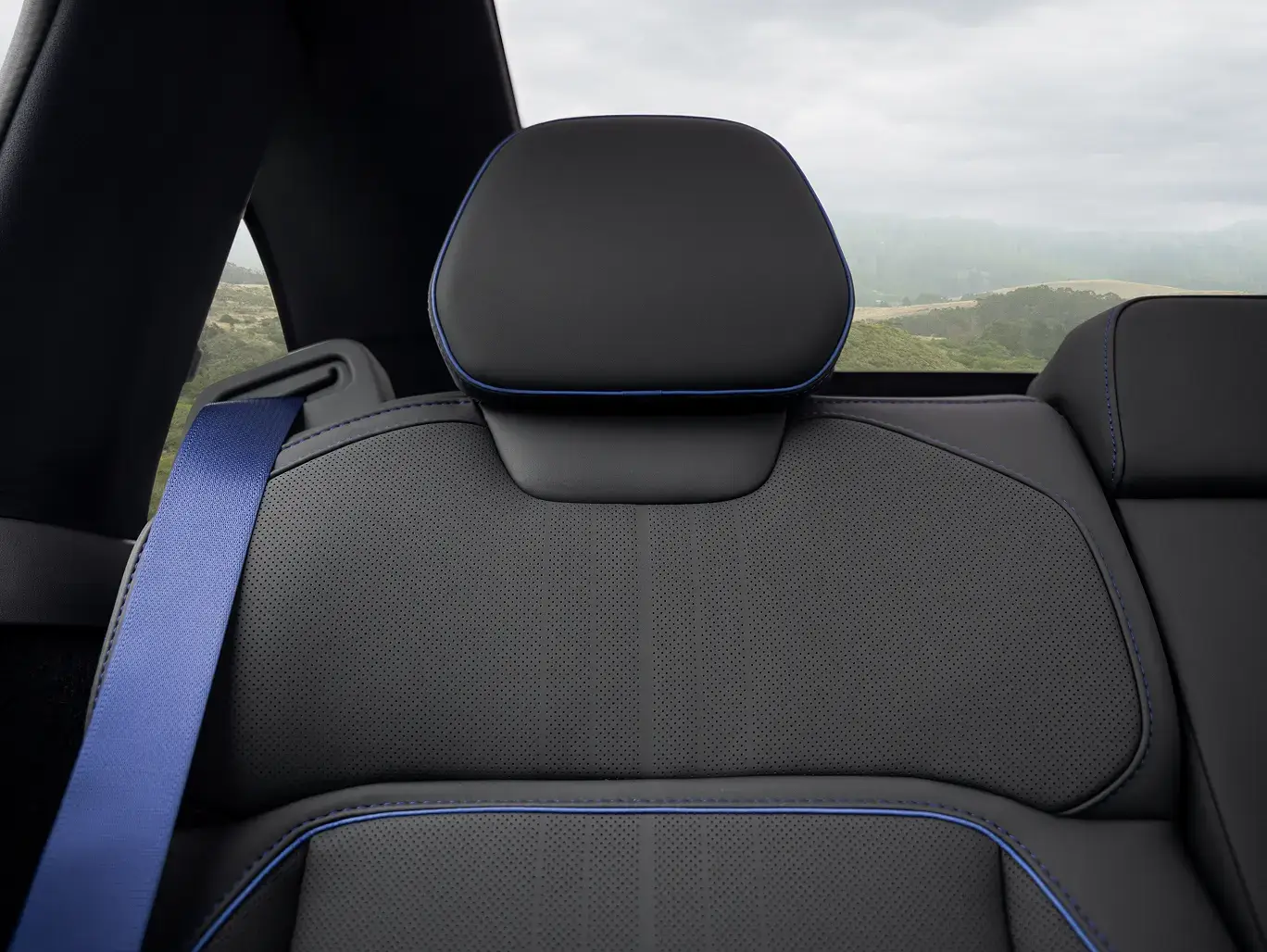Black perforated leather car seat with blue piping and headrest, blue seatbelt visible, scenic landscape through rear window.