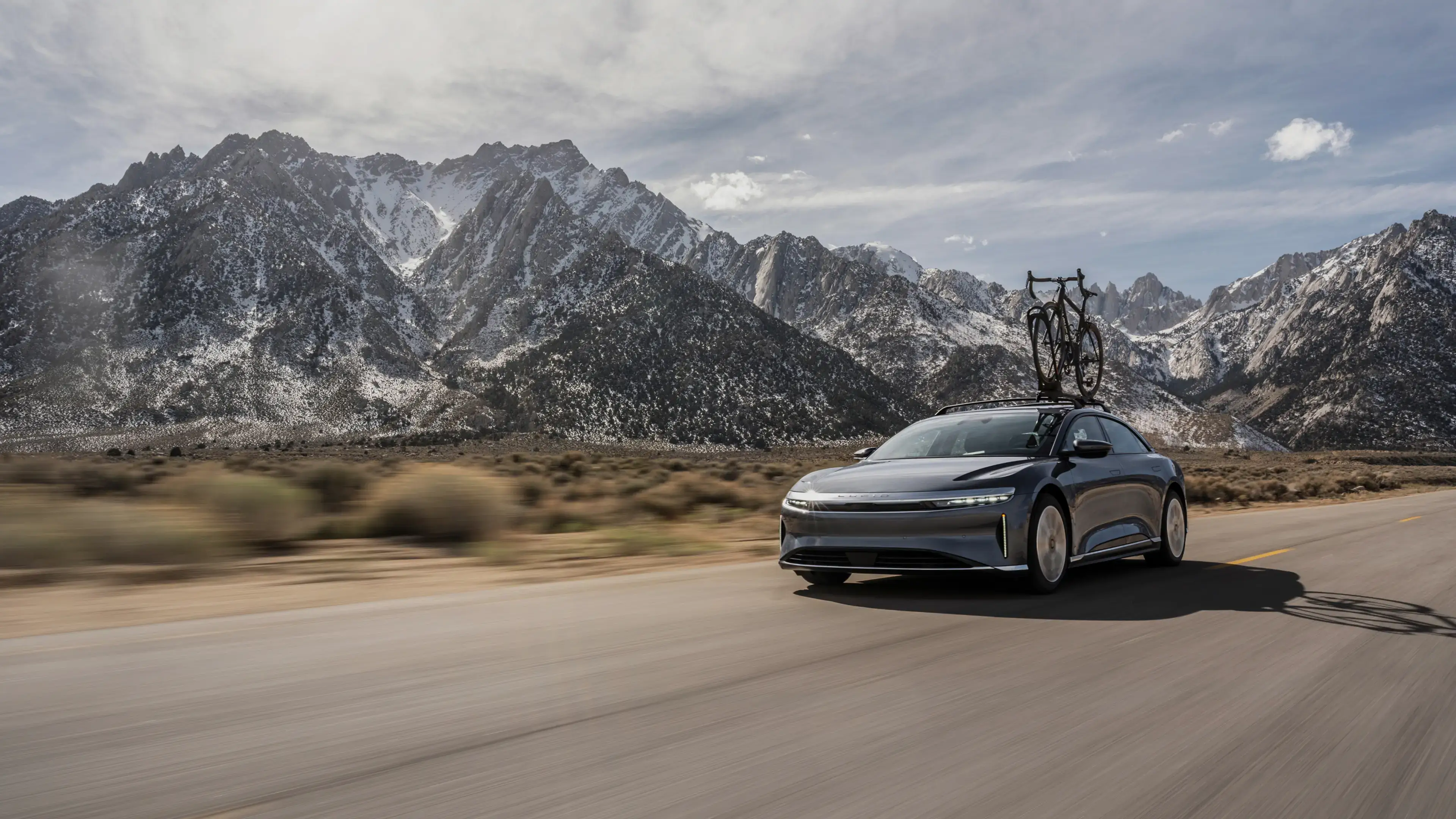 Lucid Air with bicycle on roof rack driving on open road with snow-capped mountains in background.
