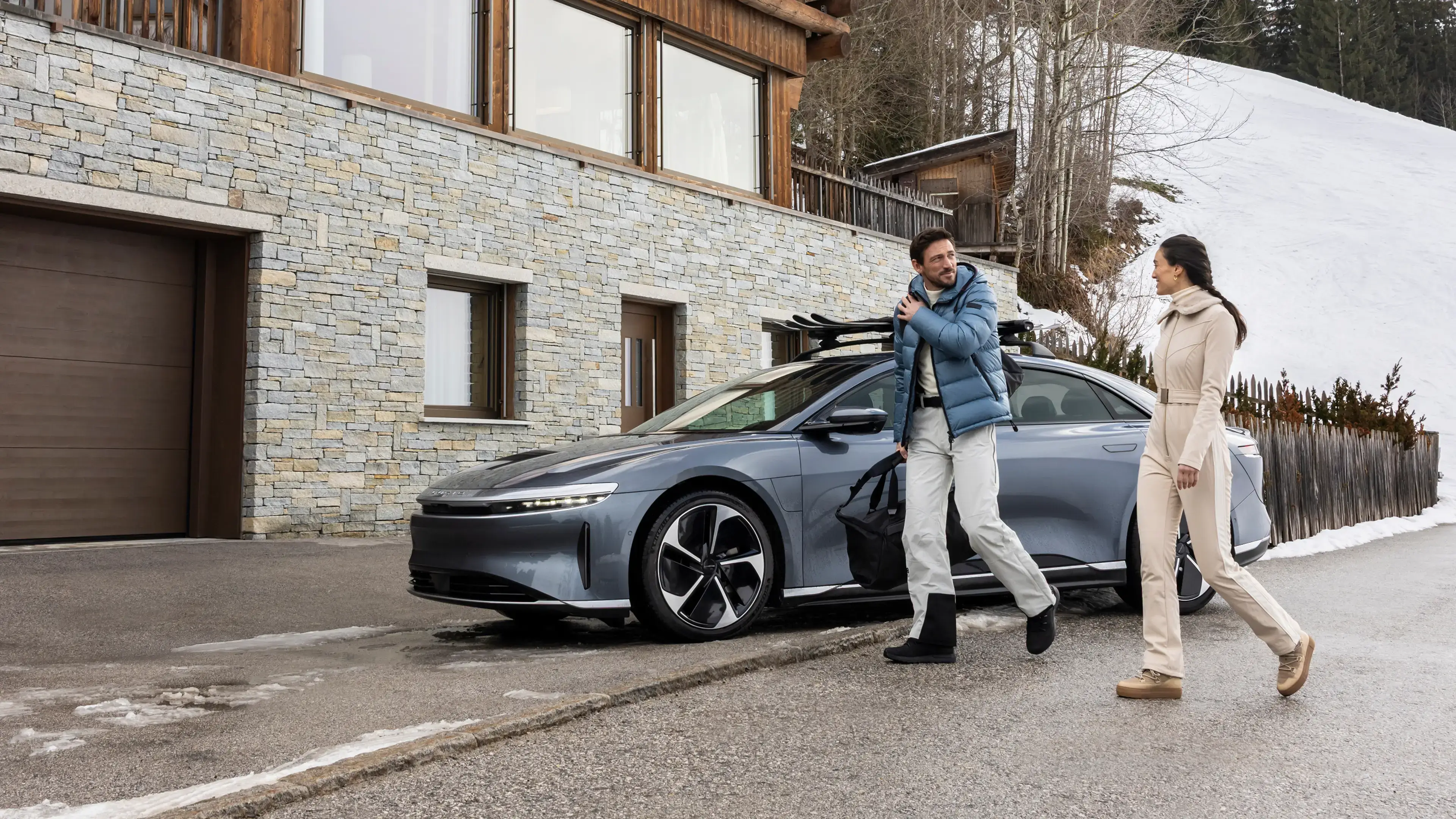 Deux personnes passent devant une Lucid Air garée devant un chalet de montagne rustique, avec de la neige en arrière-plan.