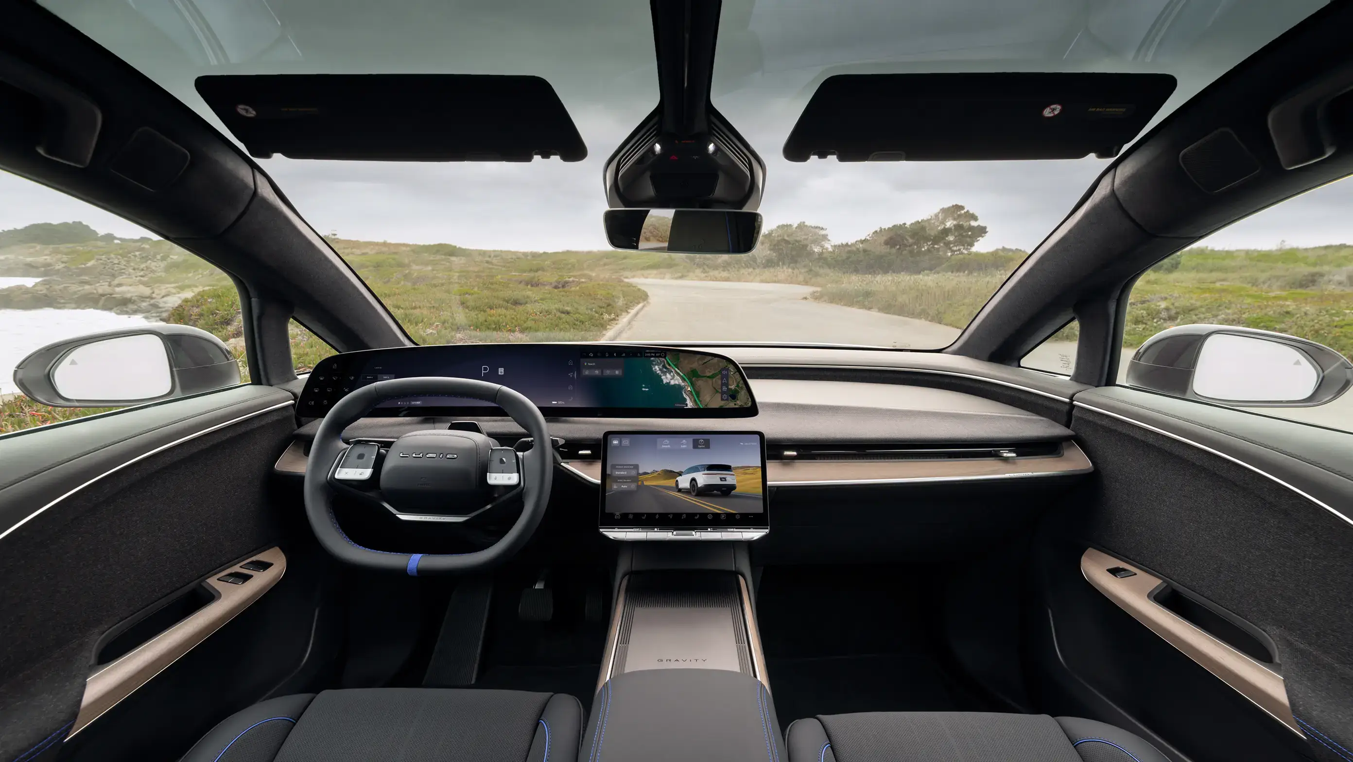 Interior of a Lucid Gravity SUV featuring a sleek dashboard, large touchscreens, panoramic windshield, and a coastal road view.