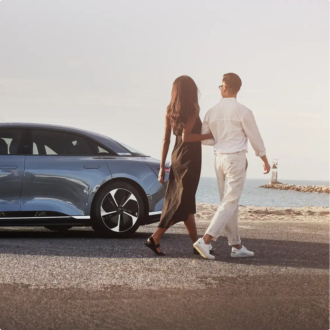A couple walks together near a blue Lucid Air car parked by a coastal road, with a lighthouse and ocean in the background.