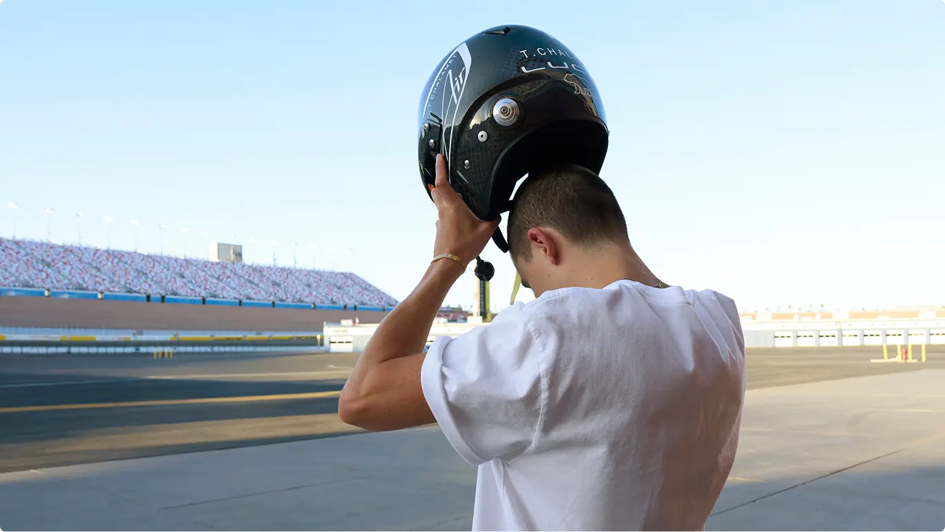 Timothée Chalamet putting on black racing helmet at a racetrack with stadium seating visible in the background under blue sky.