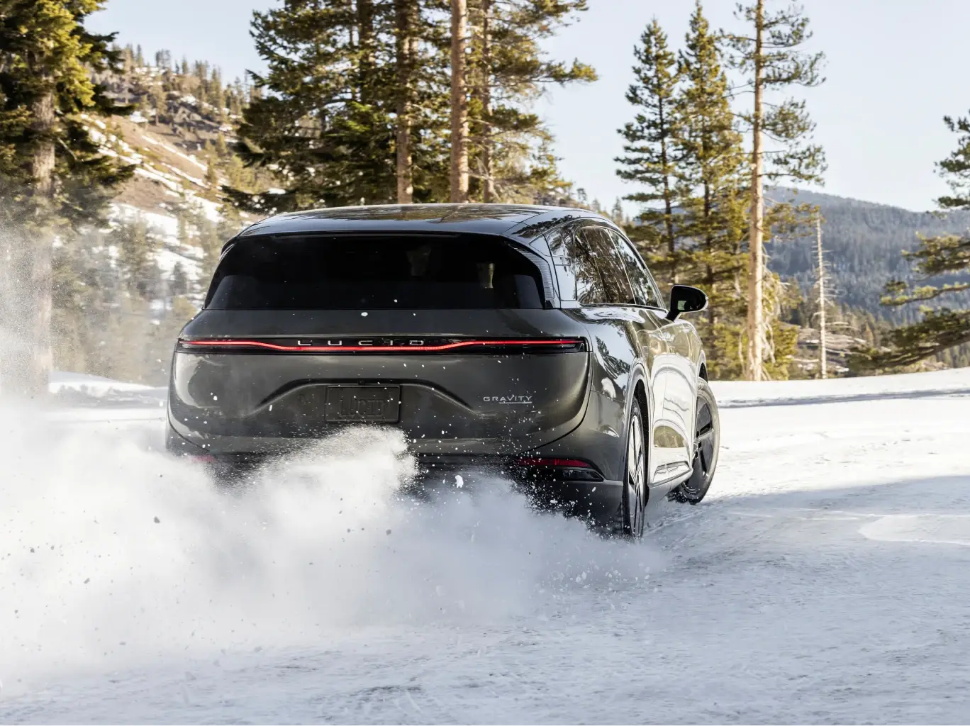 Lucid SUV driving through snowy terrain, kicking up powder against a backdrop of tall pine trees and mountains.